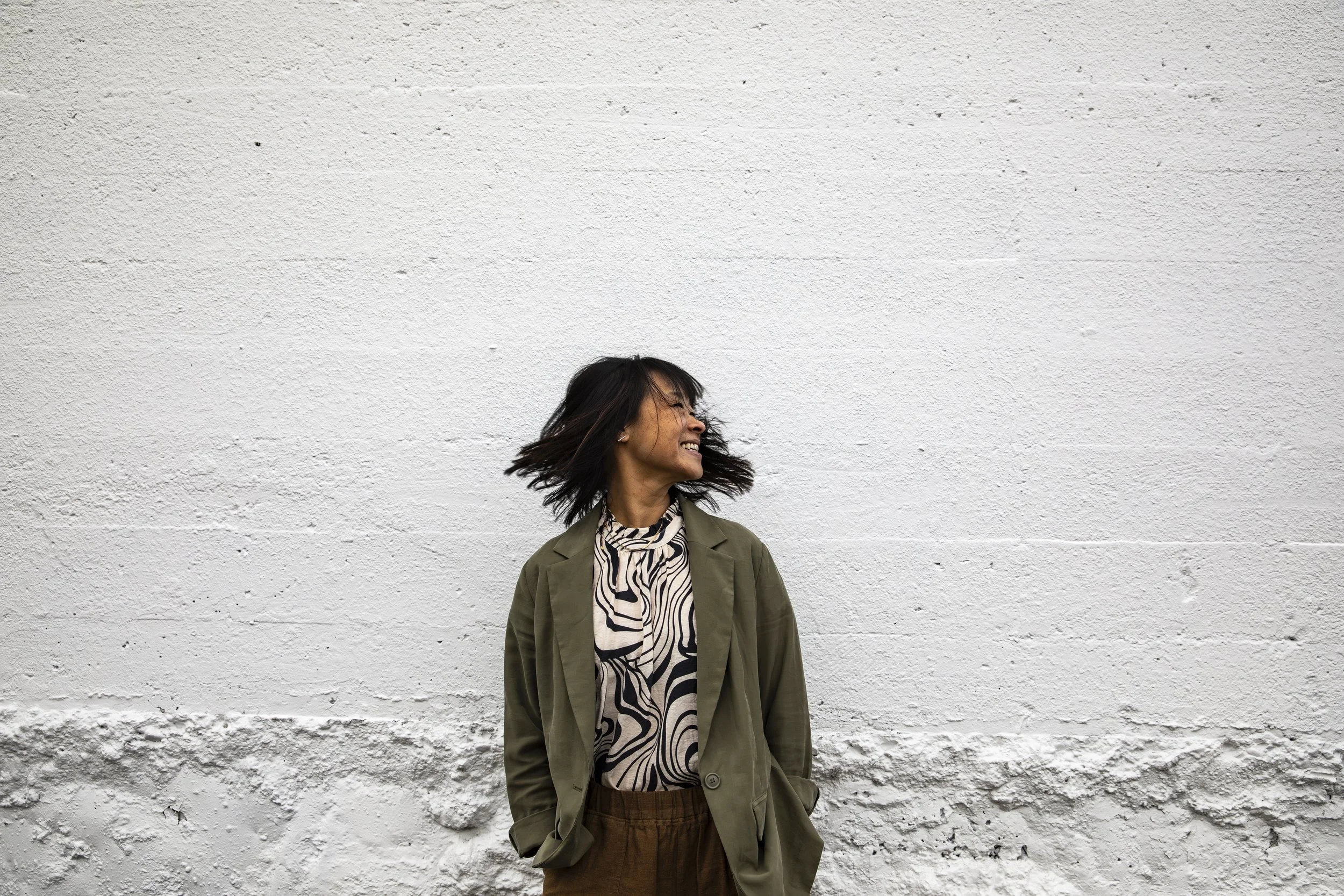 A woman with black hair is smiling and turning her head to the right while standing against a textured white wall. She is wearing a patterned blouse, an olive green jacket, and brown pants.