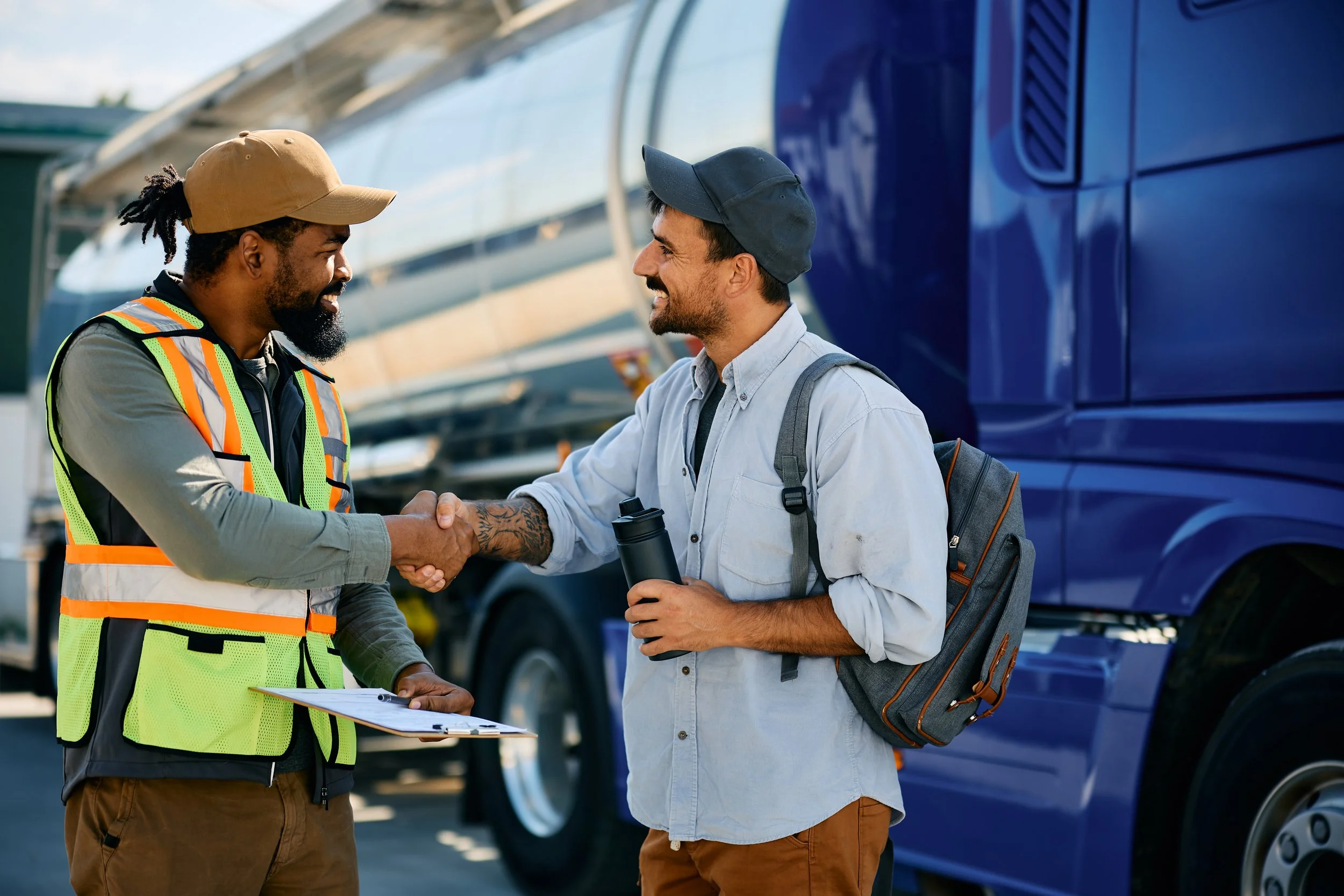 Two men shaking hands in front of a blue truck; one man is wearing a safety vest and holding a clipboard, the other is in casual clothes with a backpack holding a water bottle, both smiling.