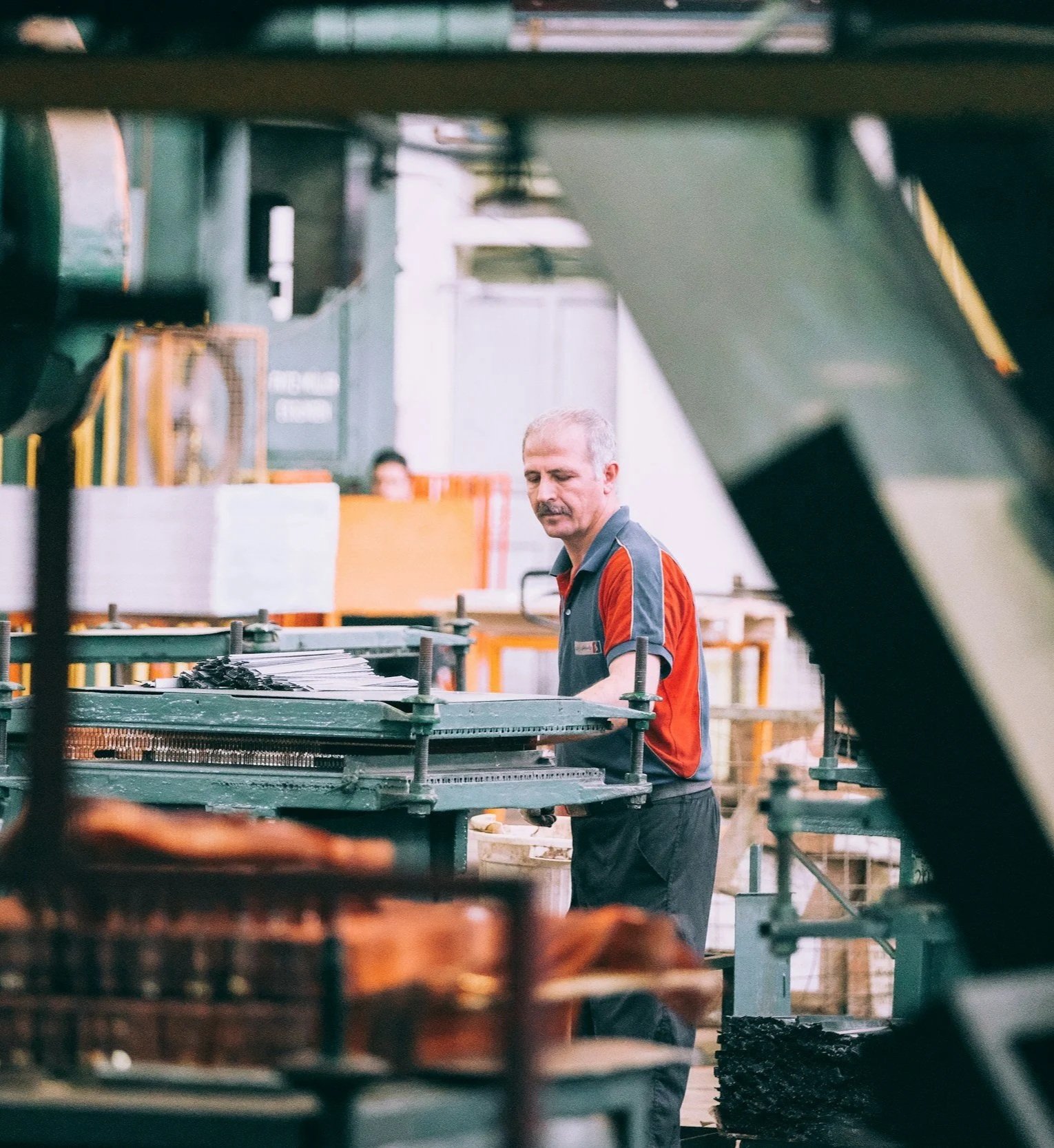 A worker in a factory, standing at a work station with machinery, dressed in a black and red work shirt, with gray hair and beard, surrounded by industrial equipment and materials.