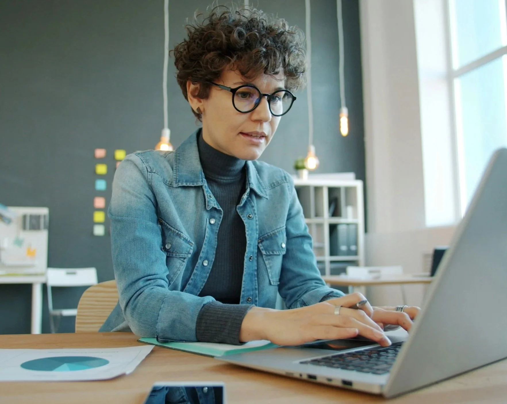 A woman with curly hair and glasses working on a laptop in an office with a dark wall and white shelving in the background.