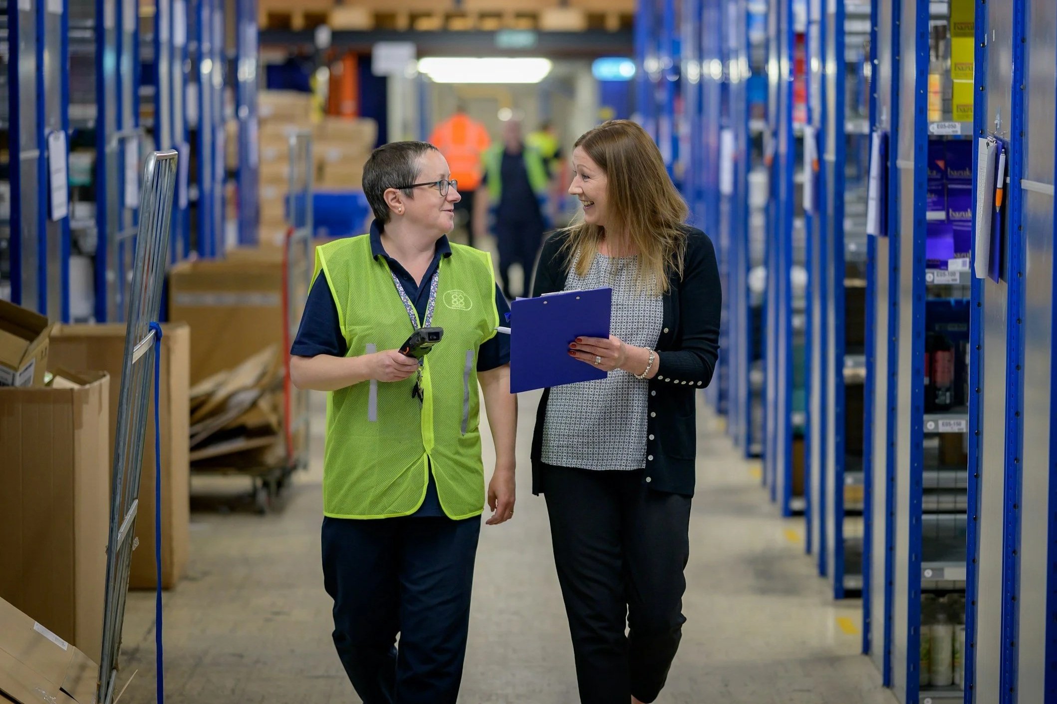 Two women walking and talking in a warehouse. One woman in a safety vest holding a scanner, the other holding a clipboard. Shelves filled with boxes and items are visible in the background.
