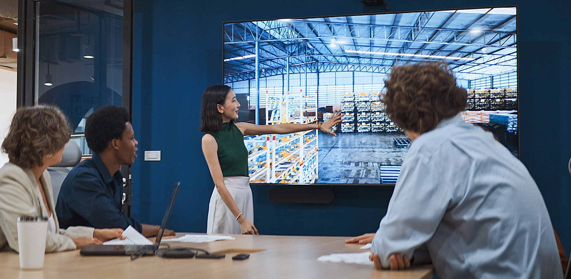 Businesswoman presenting warehouse interior on a large screen to a group of three diverse professionals in a meeting room.