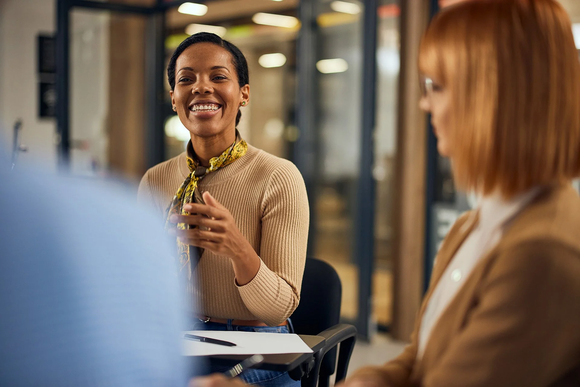 A woman with short black hair smiling and gesturing during a meeting with colleagues in an office setting.