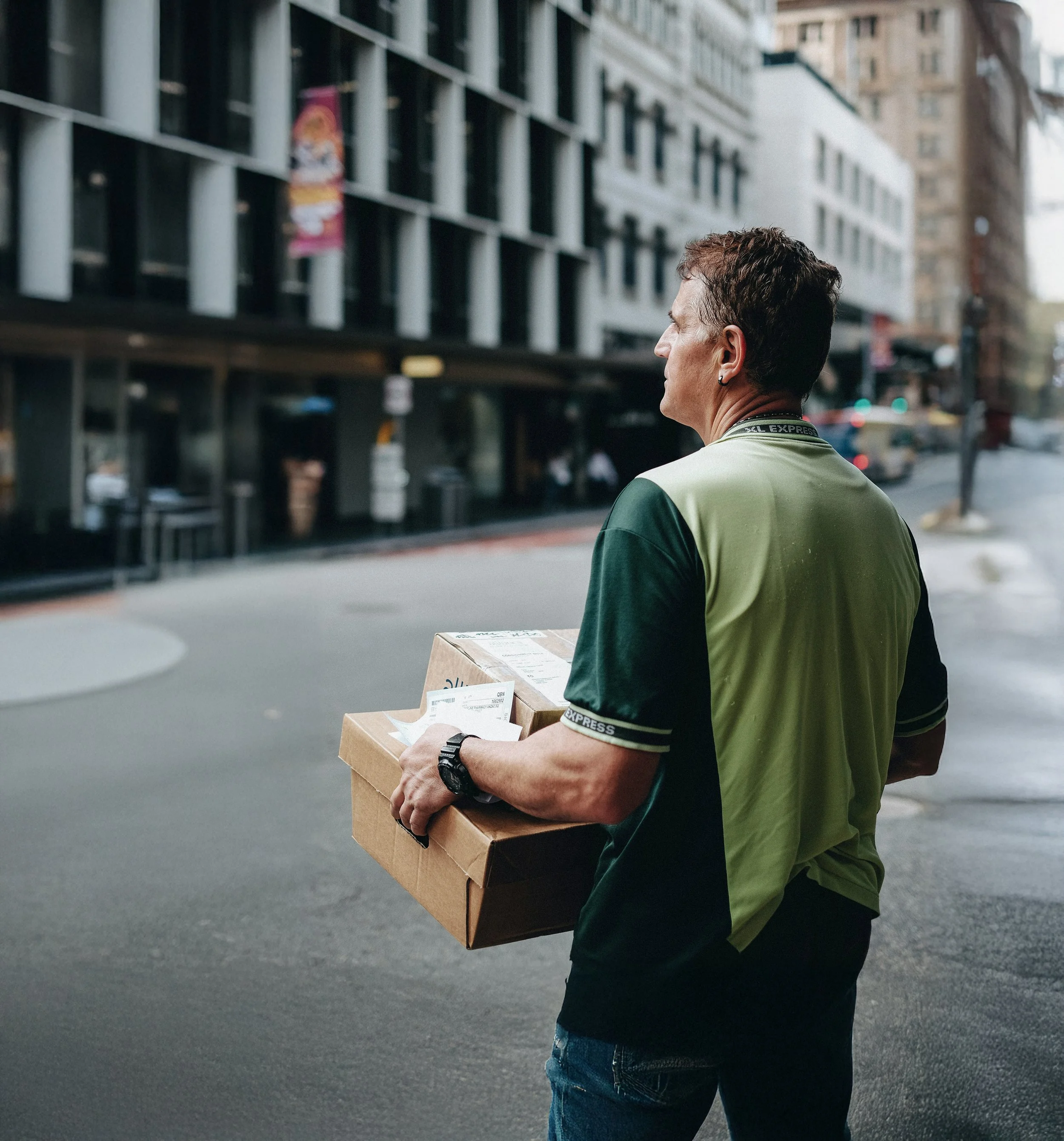 A mail carrier wearing a green and black uniform delivers a package on a city street.