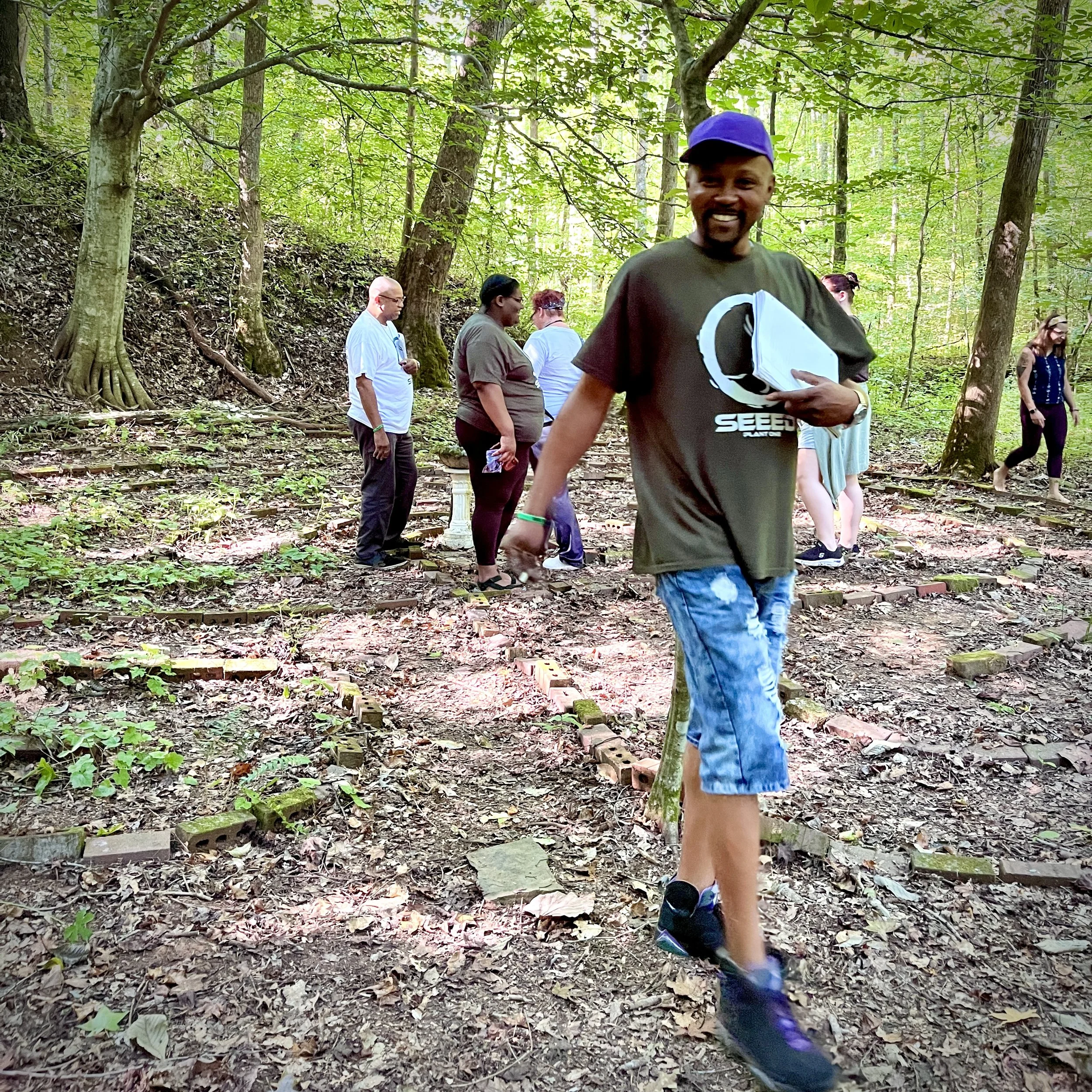 Smiling man with a purple cap and dark T-shirt holding a white object, walking in a wooded area with trees and other people in the background.