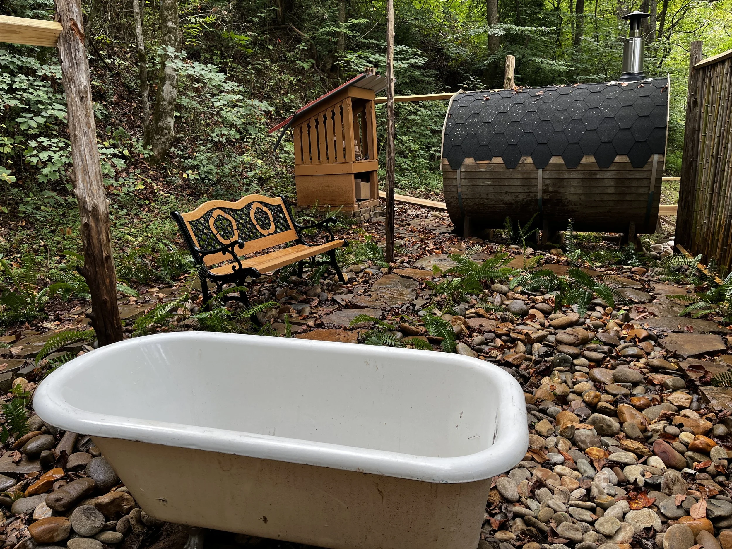 A white bathtub in a Forest spa wooded outdoor setting with rocks and ferns, a wooden bench, and a cedar barrel sauna.