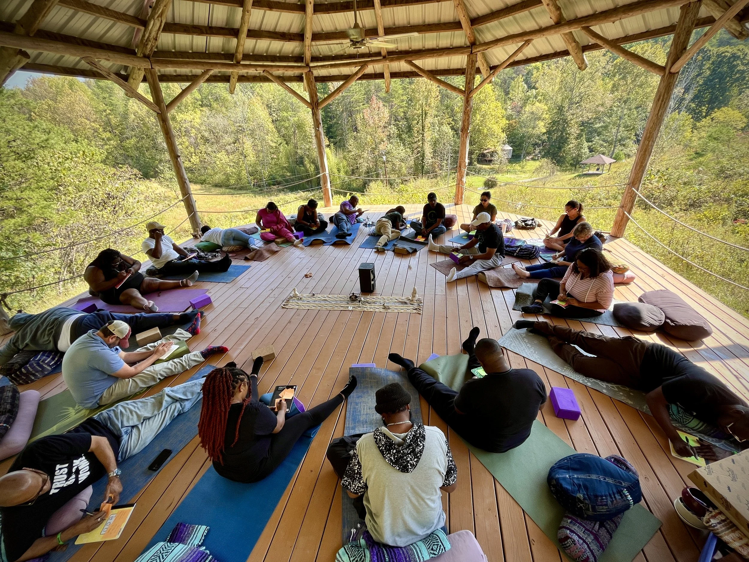 Group of people practicing yoga on mats outdoors on a wooden deck, surrounded by trees.