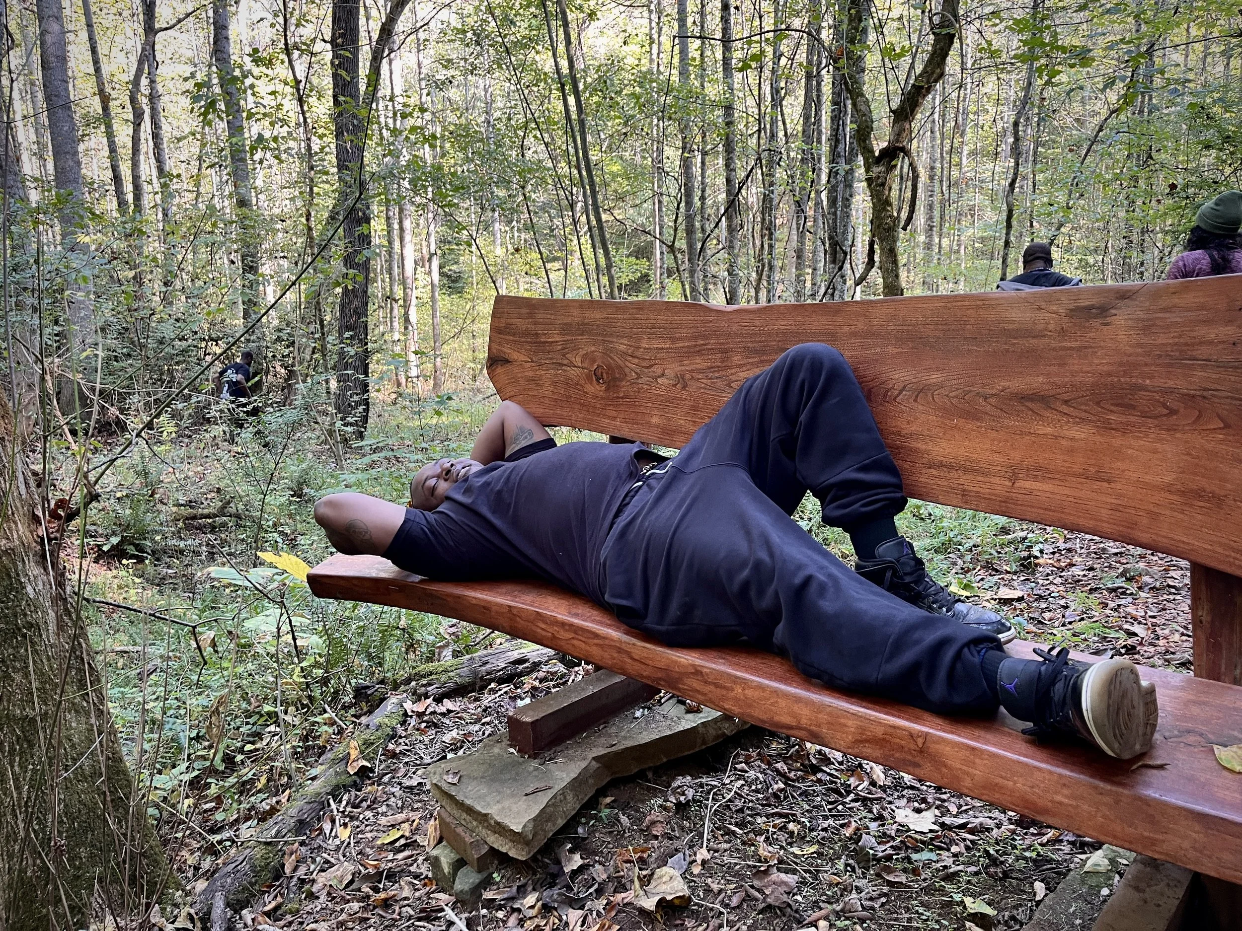 A person lying on a wooden bench in a forest, with their eyes closed and arms positioned behind their head, surrounded by trees and foliage.