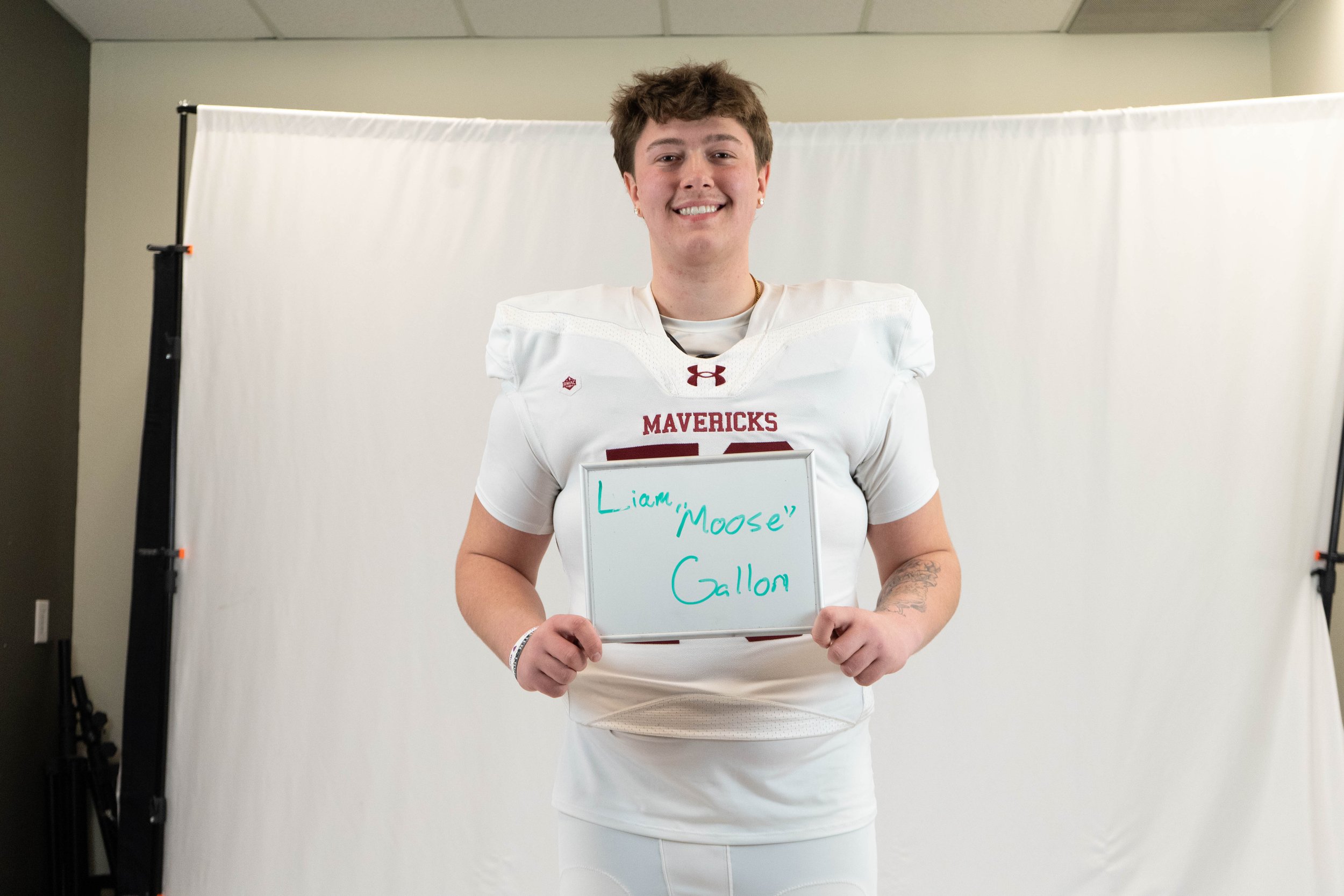 Young woman in a white football uniform holding a whiteboard that says "Liam 'Moose' Gallon" in front of a plain white background.