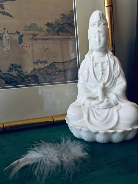 White porcelain or ceramic figurine of a seated goddess or spiritual figure with a headdress, placed on a green surface with a feather nearby, and framed artwork or scroll in the background.