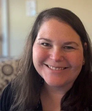 A woman with long dark hair smiling at the camera in an indoor setting.