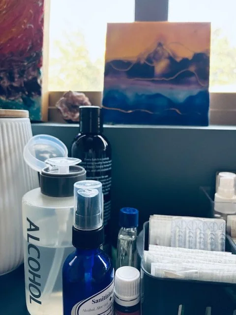 Various bottles, including alcohol, hand sanitizer, and other toiletries, on a clinic countertop with a window and landscape painting in the background.