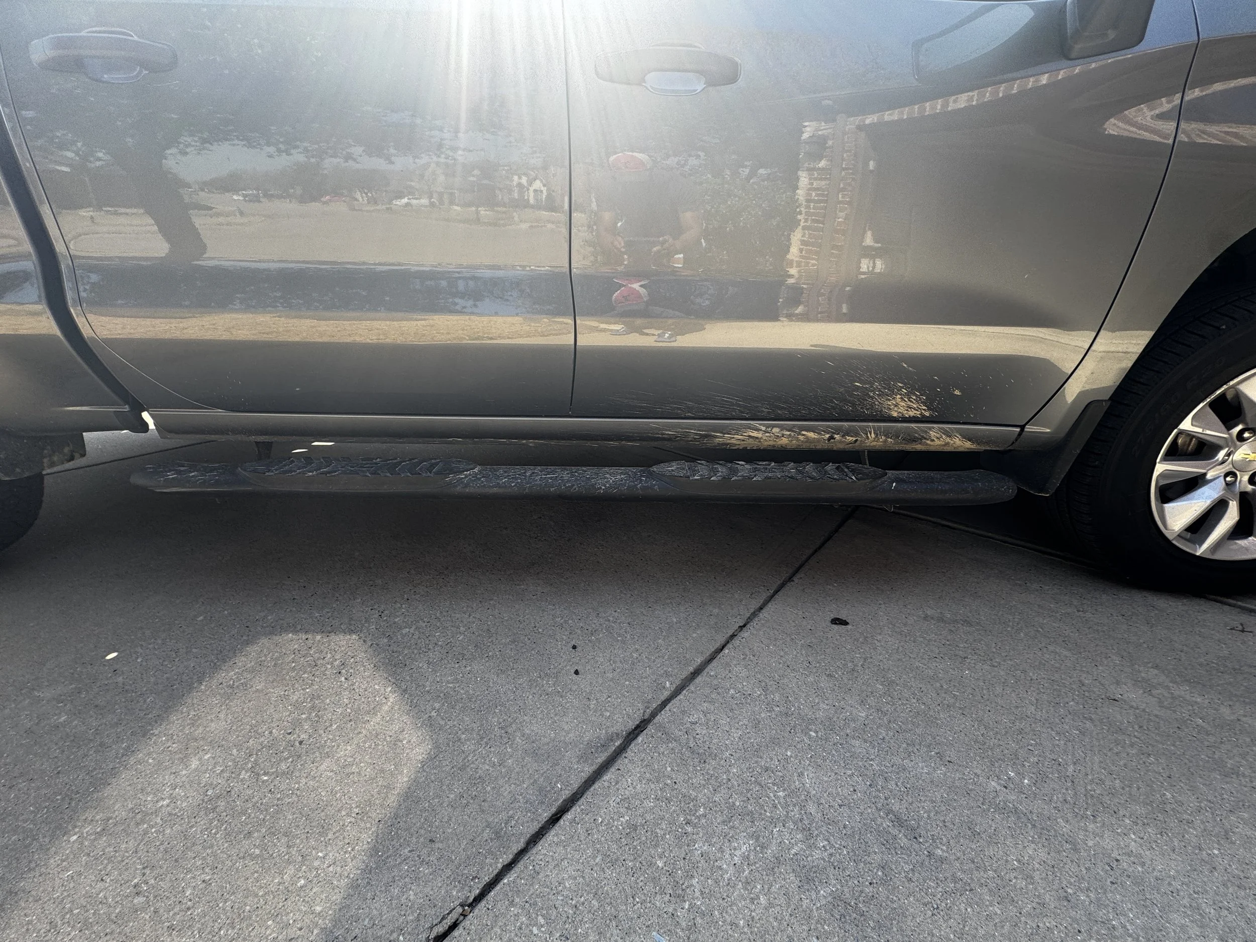 A silver vehicle with damage on the side, including dirt and scratches on the lower part of the door and side step, parked on a concrete driveway.