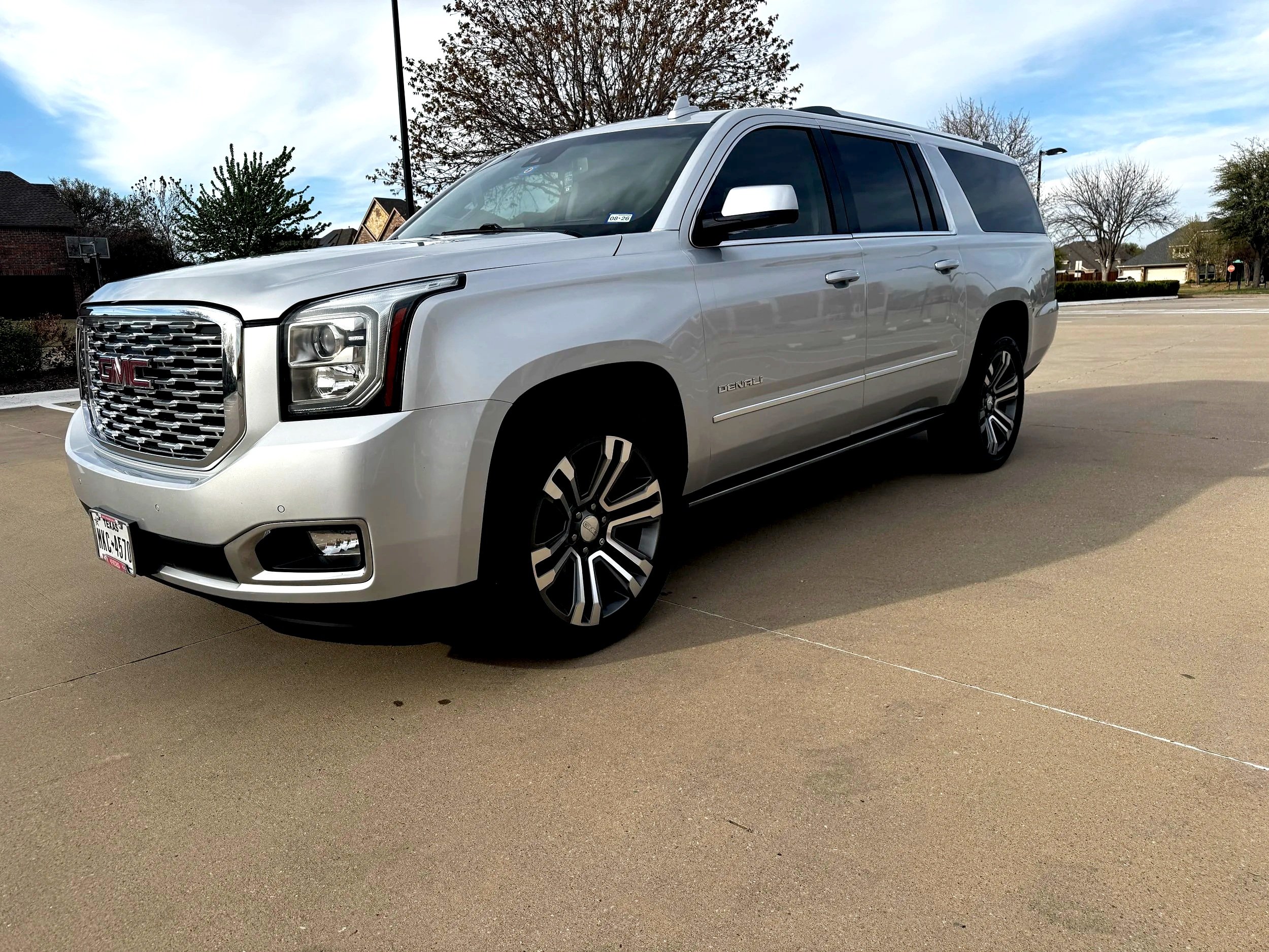 A silver GMC Yukon SUV parked on a concrete driveway with trees and houses in the background under a partly cloudy sky.