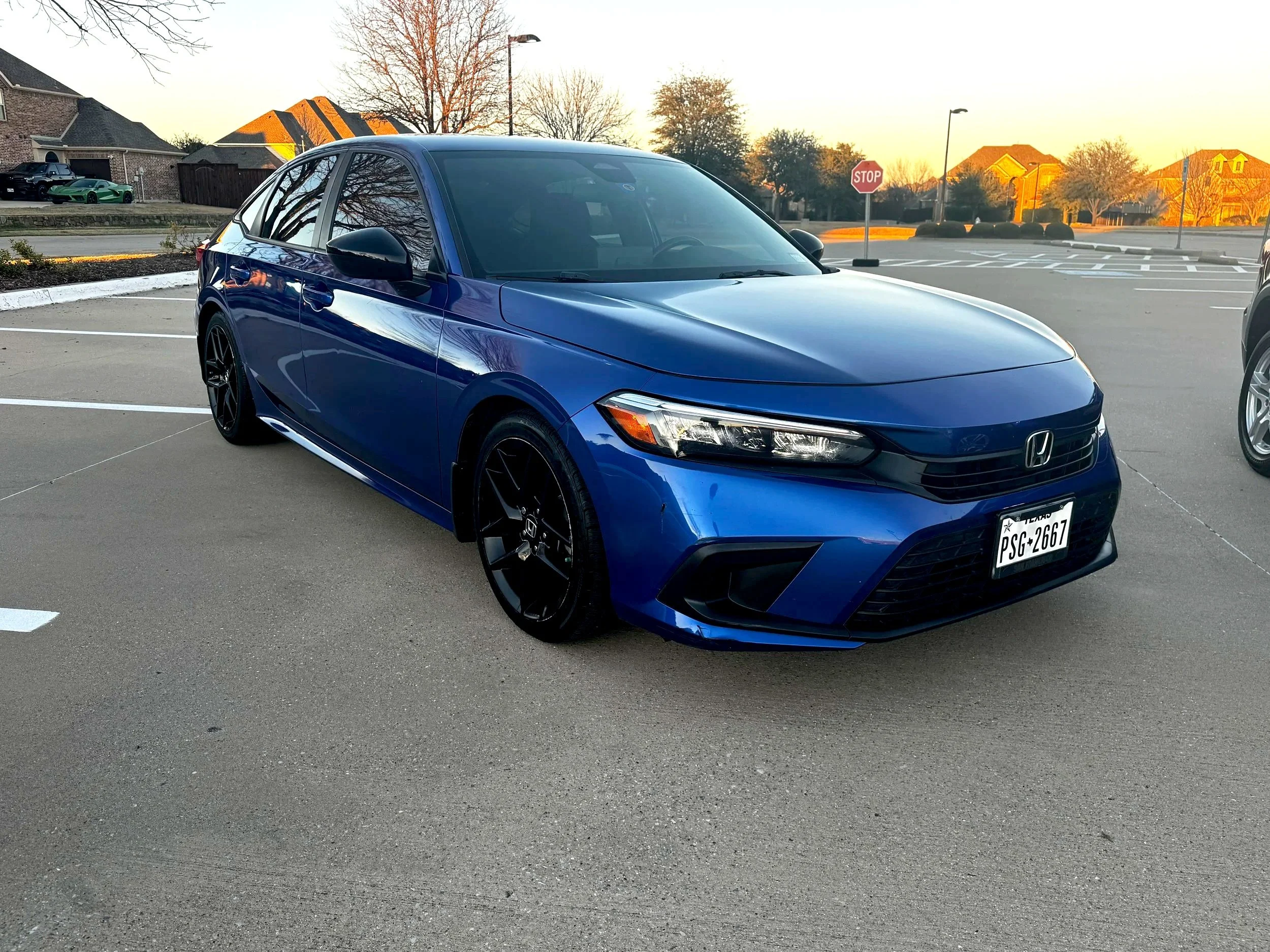 Blue Honda sedan parked in a parking lot at sunset with houses and trees in the background.