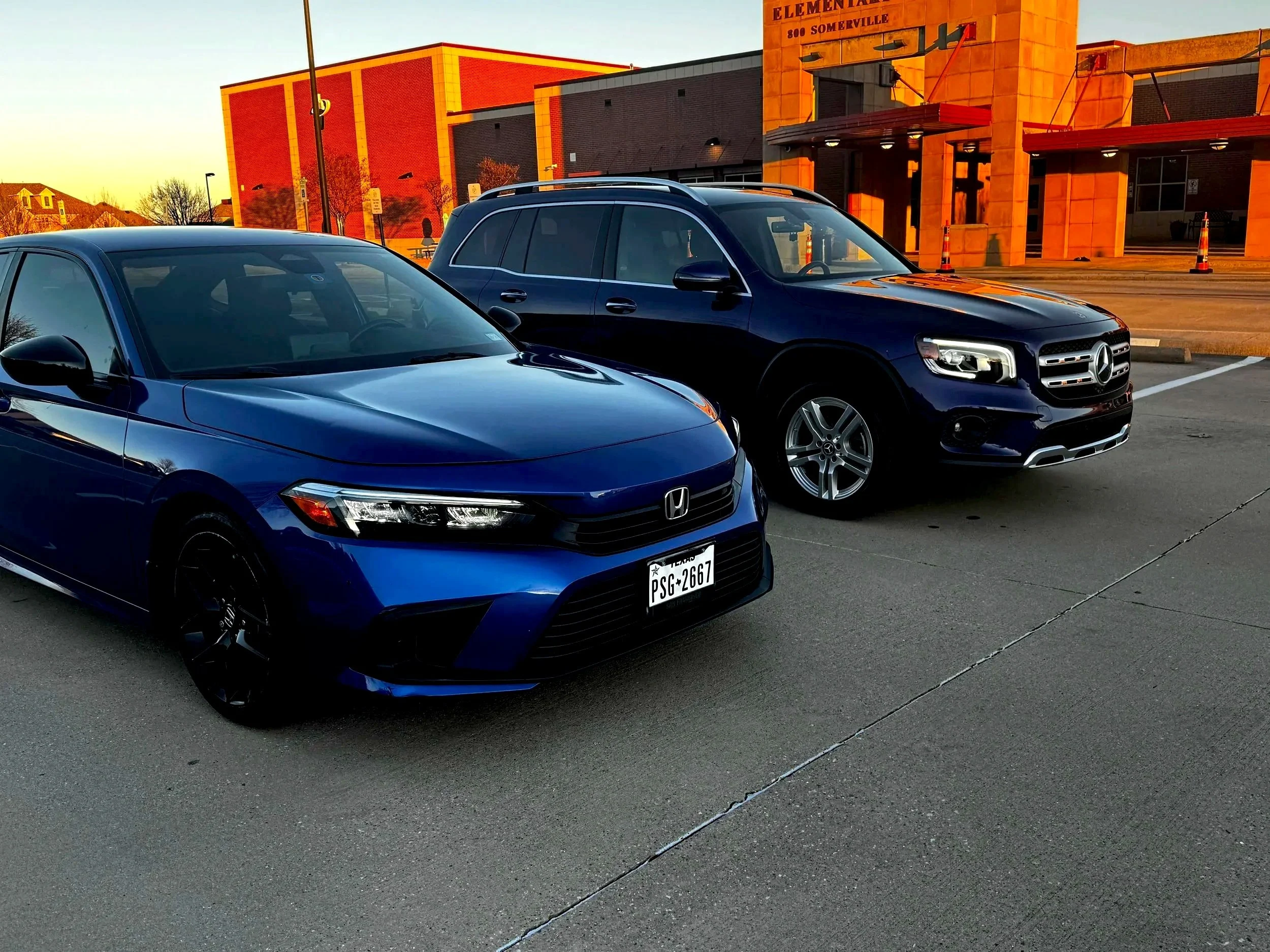 A blue Honda sedan and a dark blue Mercedes-Benz SUV parked side by side in a parking lot near a school building during sunset.