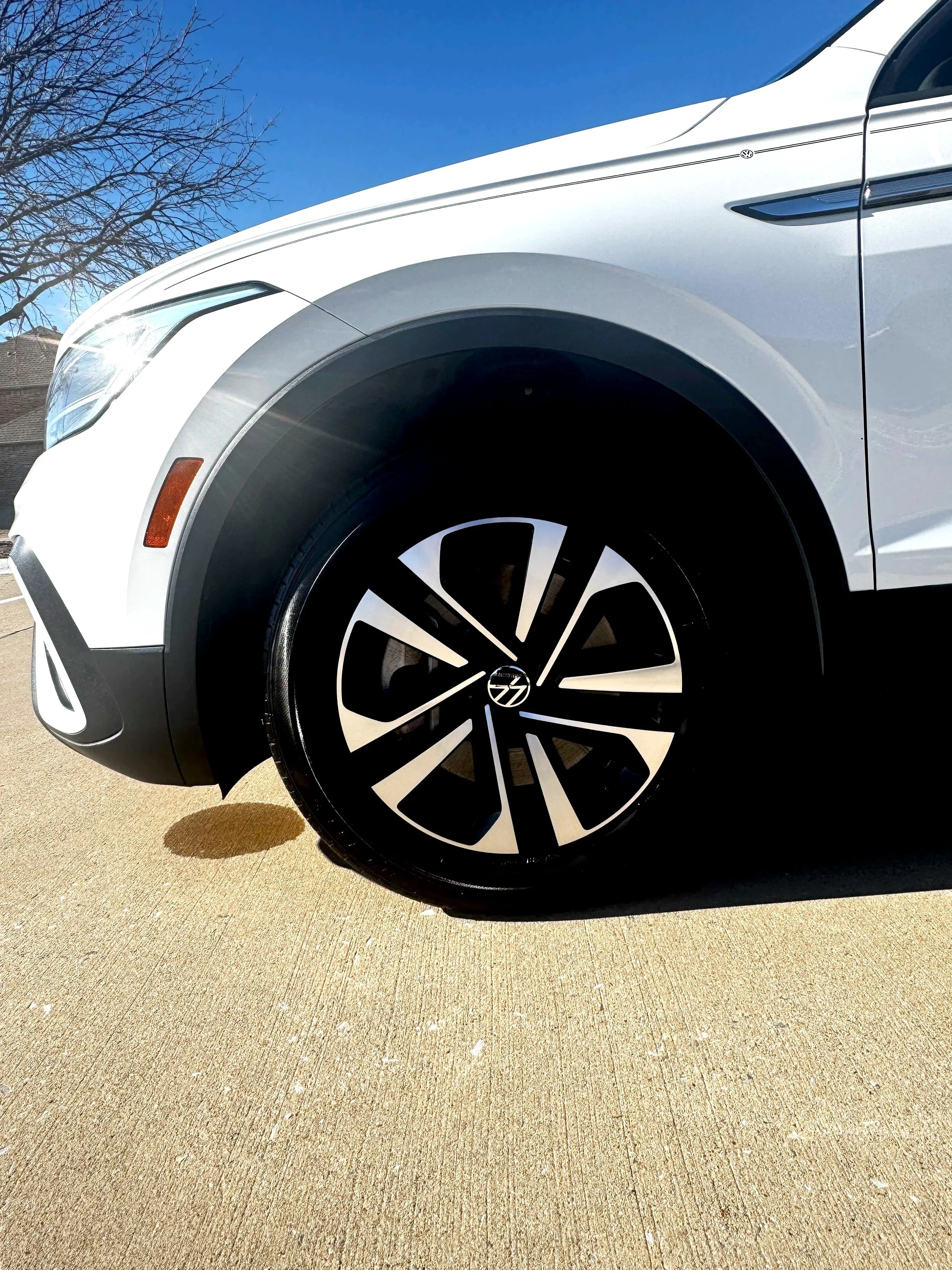 Close-up of the front left side of a white Volkswagen SUV showing the wheel and part of the front bumper against a clear blue sky and a concrete driveway.