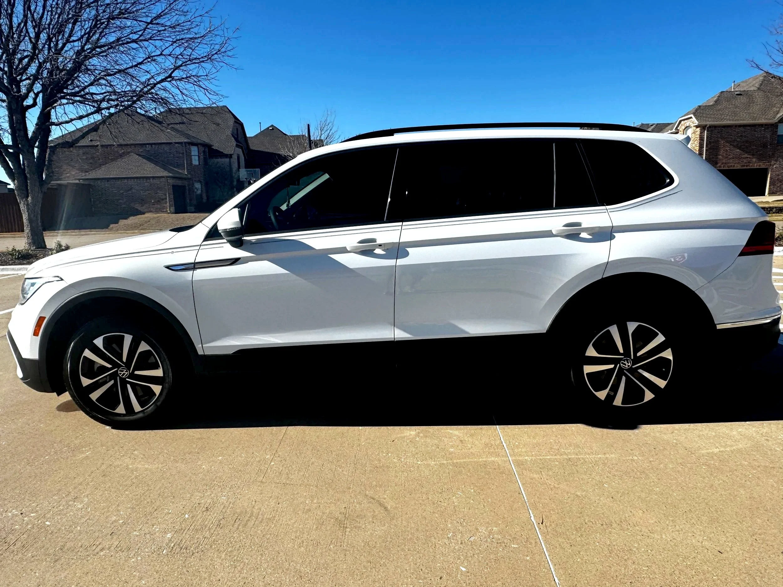 Side view of a white Volkswagen SUV parked on a driveway in a suburban neighborhood with houses and a tree in the background.