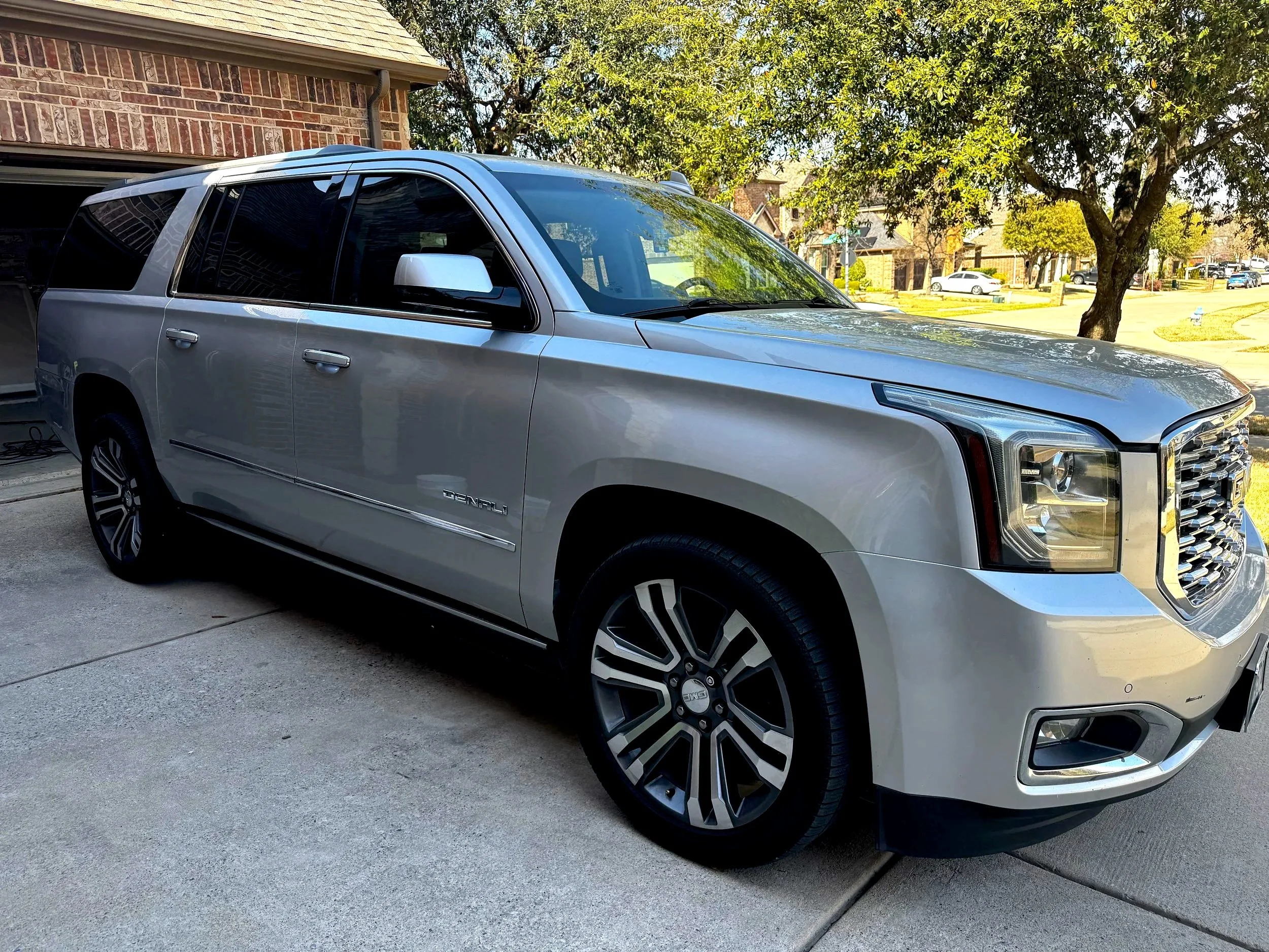 Silver GMC Yukon SUV parked in driveway with brick house and trees in background.