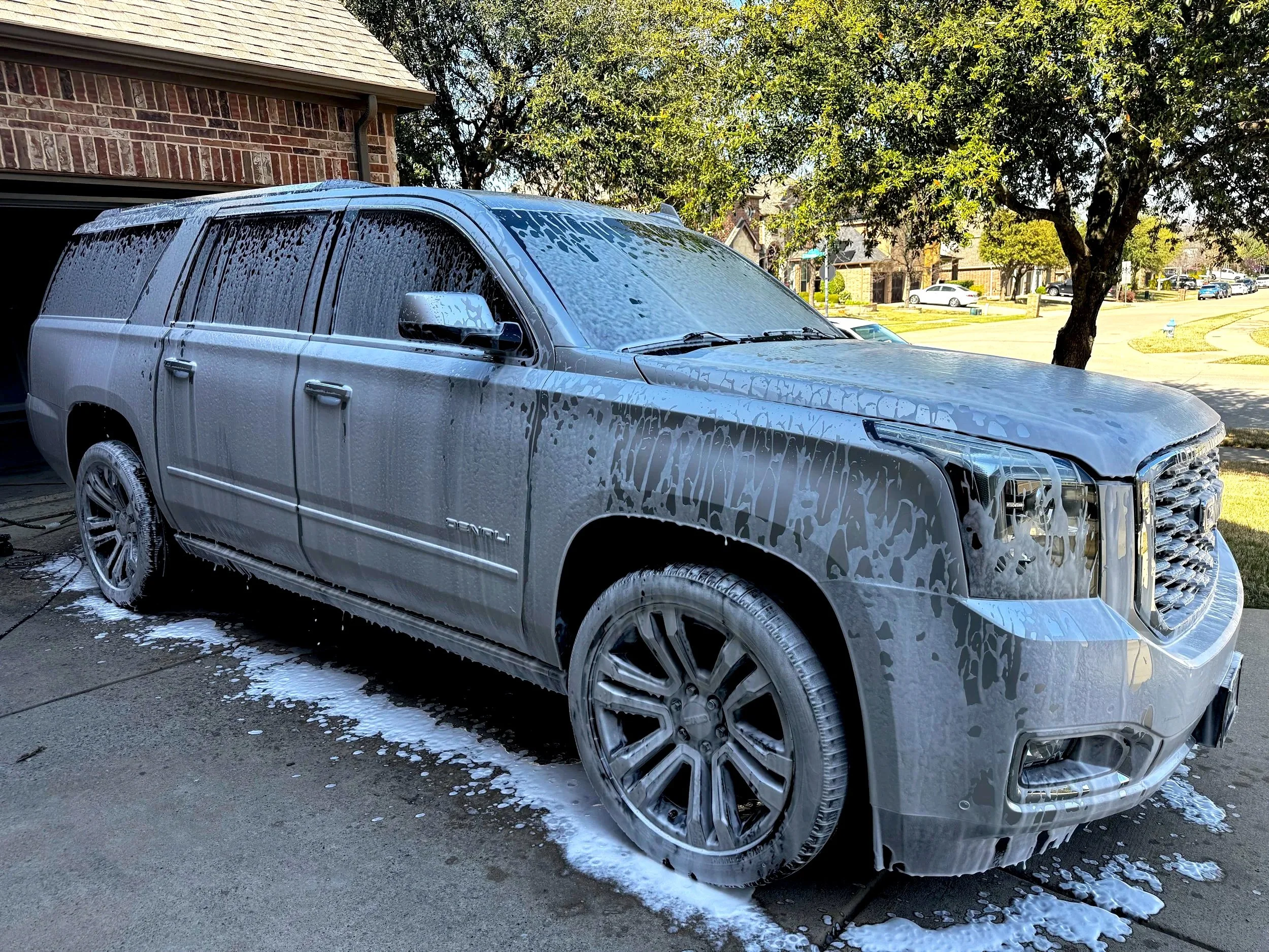 A silver SUV covered in soap suds during a car wash in a driveway.