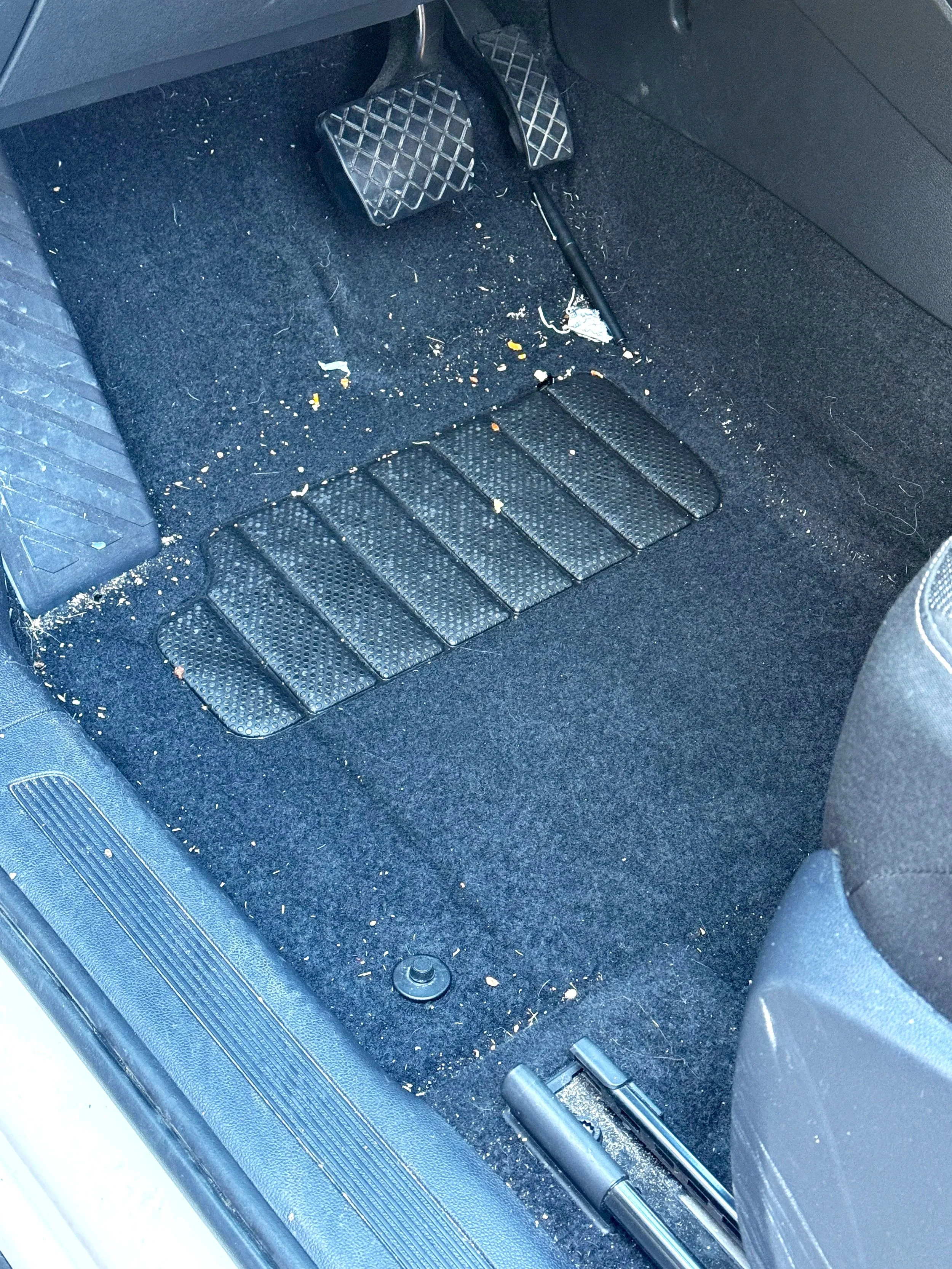 Car interior with dirty black floor mat, scattered crumbs and debris, accelerator and brake pedals, and part of passenger seat.