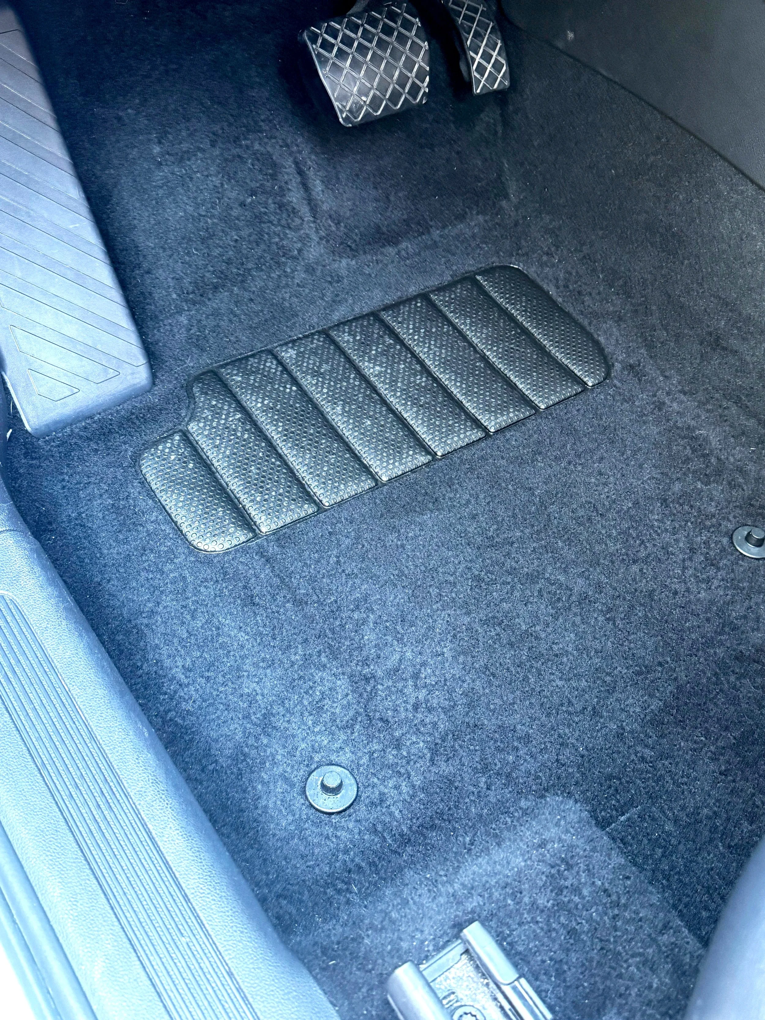 Car interior showing the black carpeted floor, accelerator and brake pedals, and a textured rubber footrest.