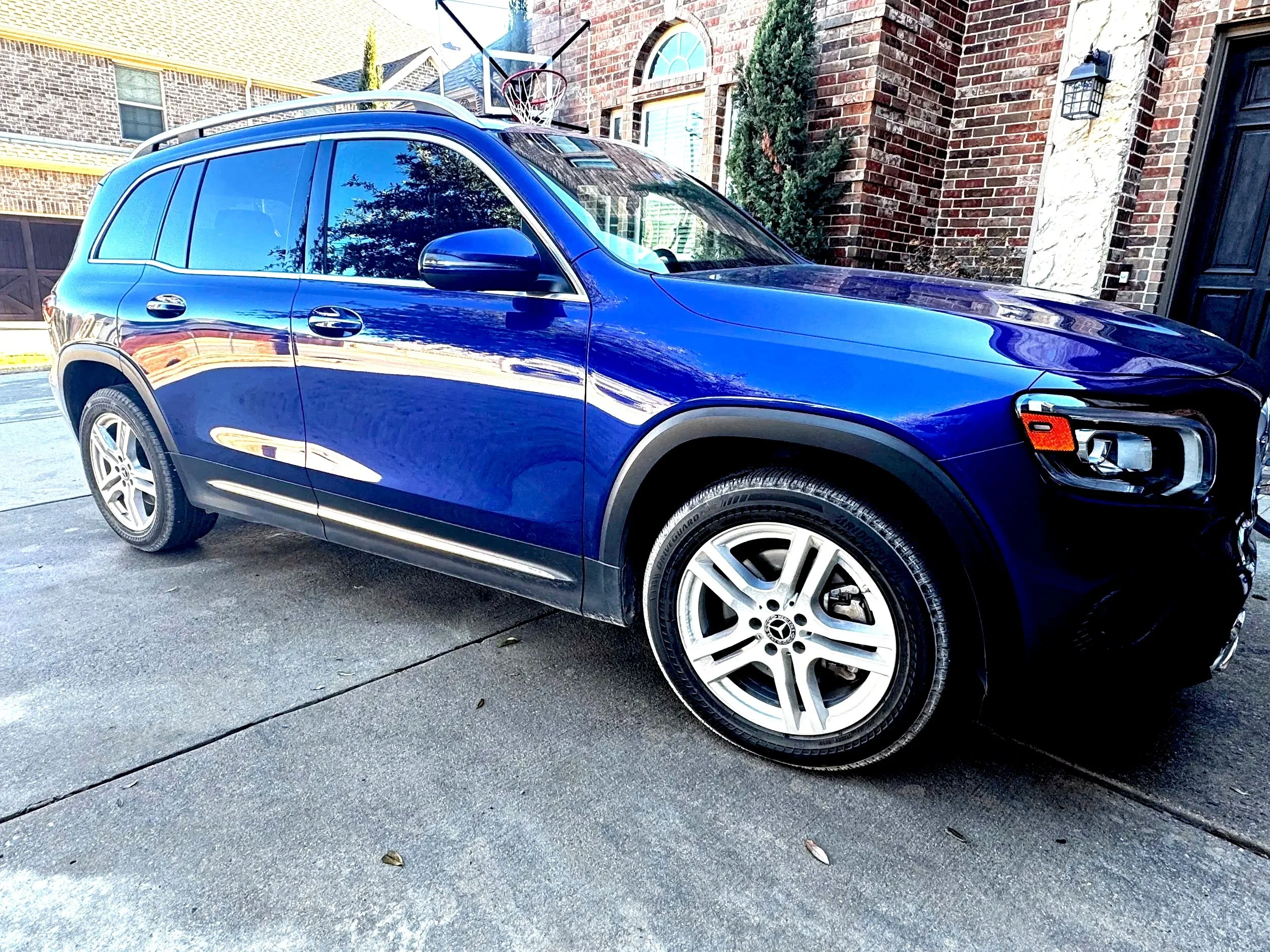 A blue Mercedes-Benz SUV parked on a concrete driveway in front of a brick house. The house has a black door and a basketball hoop mounted above it.