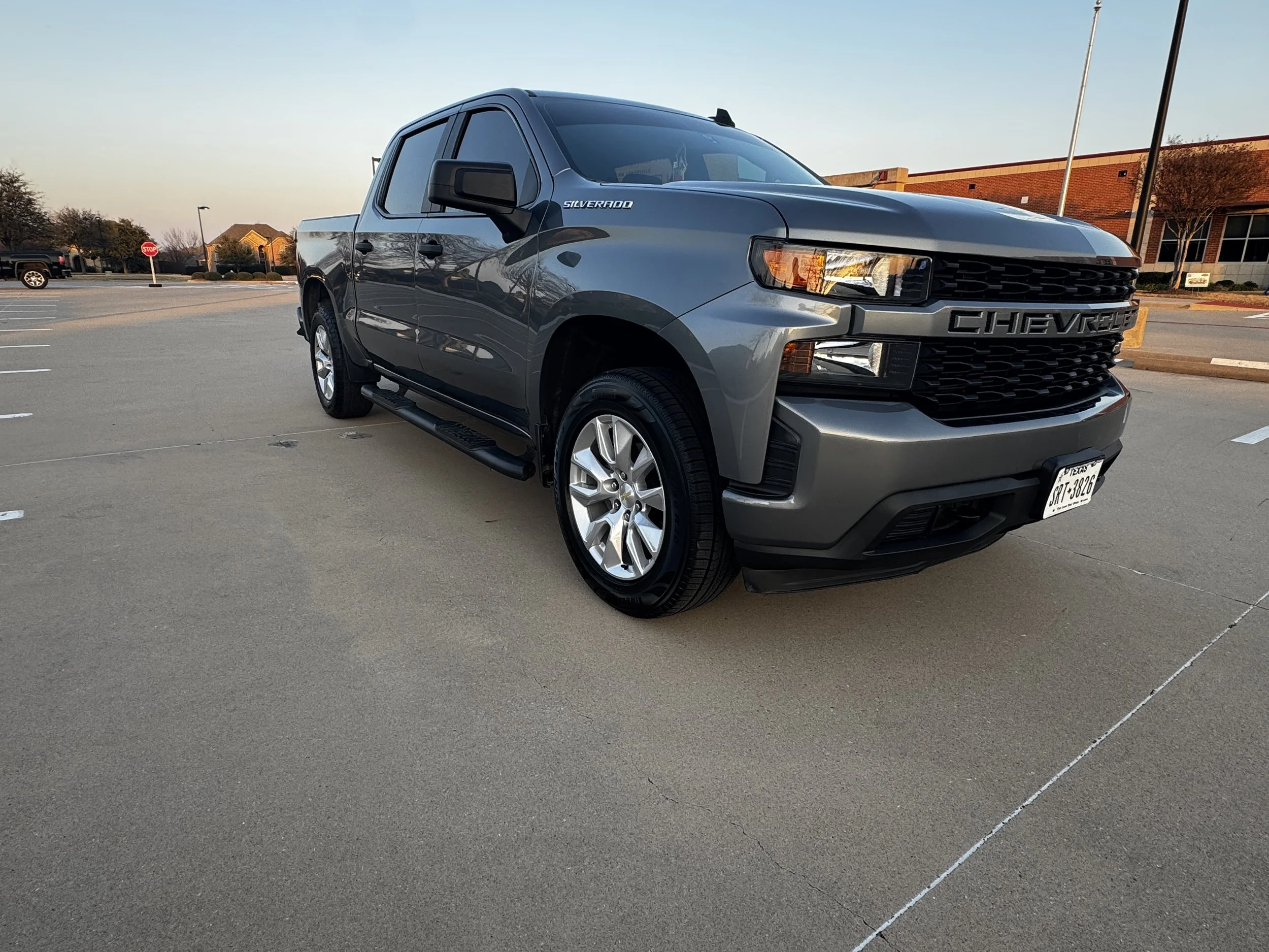 Gray Chevrolet Silverado pickup truck parked in an empty parking lot with a commercial building and trees in the background, during sunset.