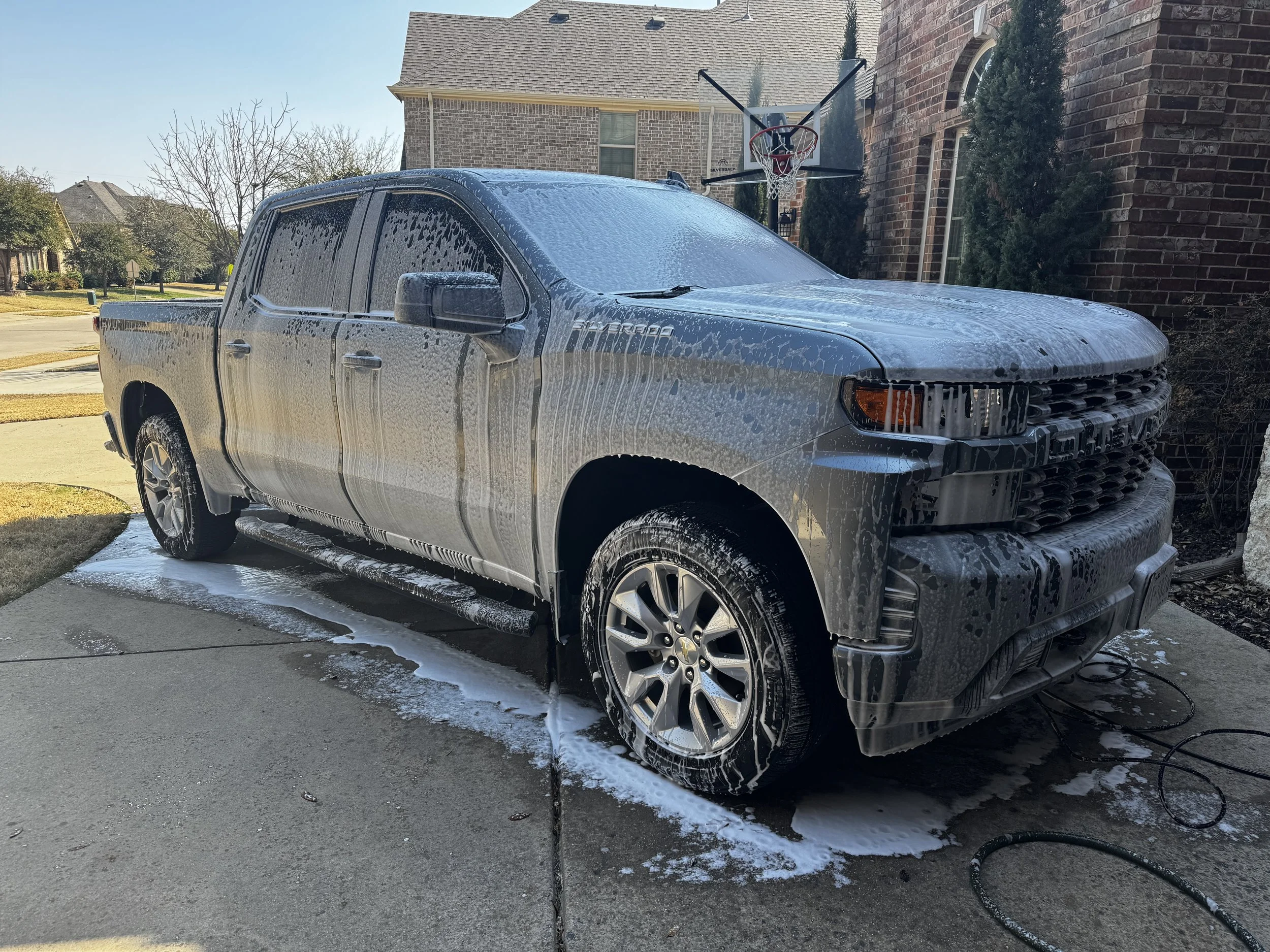 A gray pickup truck covered in soap suds during a cleaning in a driveway, with a basketball hoop in the background.