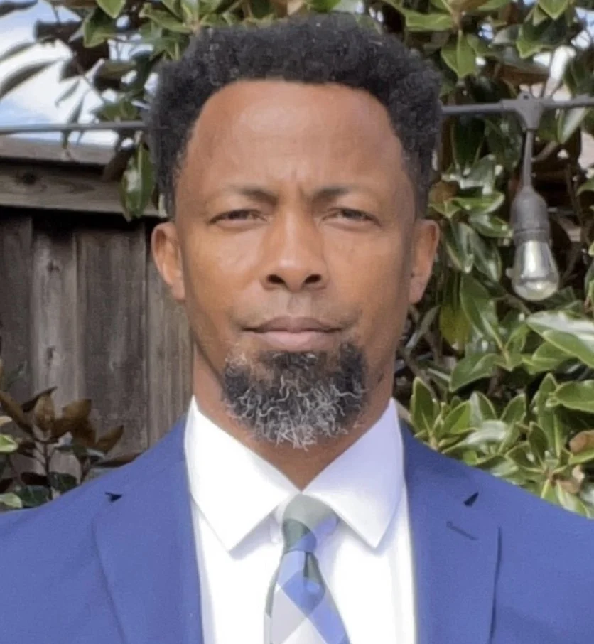 A man with dark skin, short curly hair, and a salt-and-pepper goatee, wearing a blue suit, white shirt, and a striped tie, standing in front of a leafy background with a wooden fence and outdoor string lights.