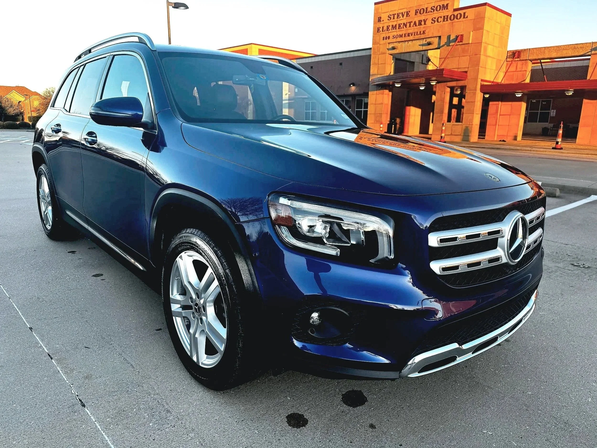 A dark blue Mercedes-Benz SUV parked in a parking lot in front of a red brick building labeled R. Steve Folsom Elementary School.