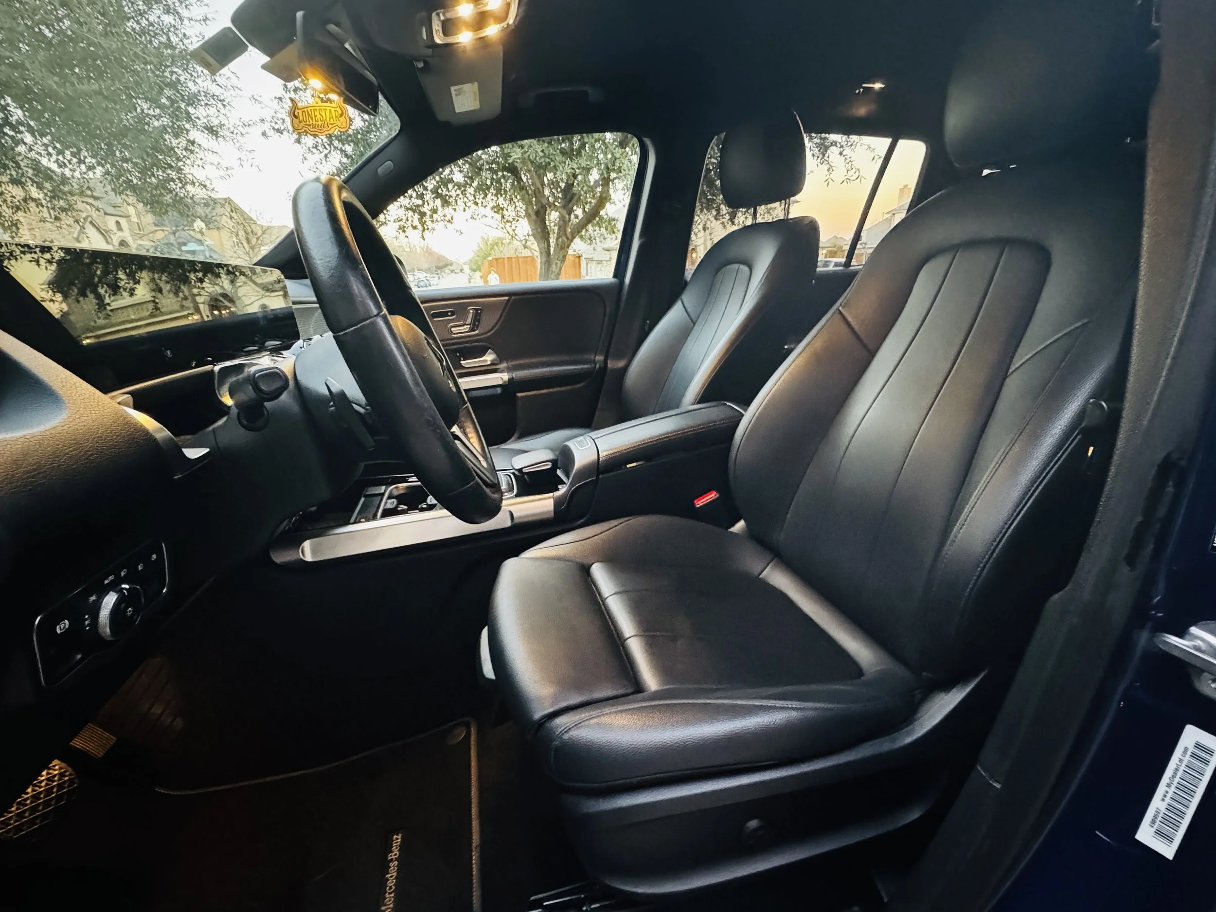 Interior of a vehicle showing two front leather seats, steering wheel, dashboard, and side window with a tree and houses outside at sunset.