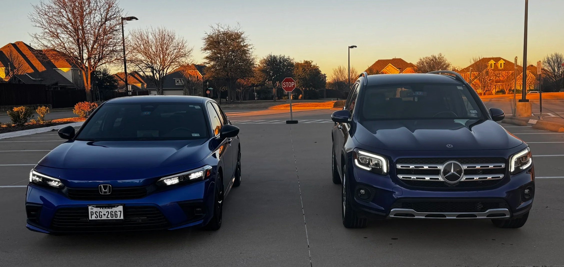 A blue Honda Civic and a blue Mercedes-Benz SUV parked side by side in a parking lot at sunset, with houses and trees in the background.