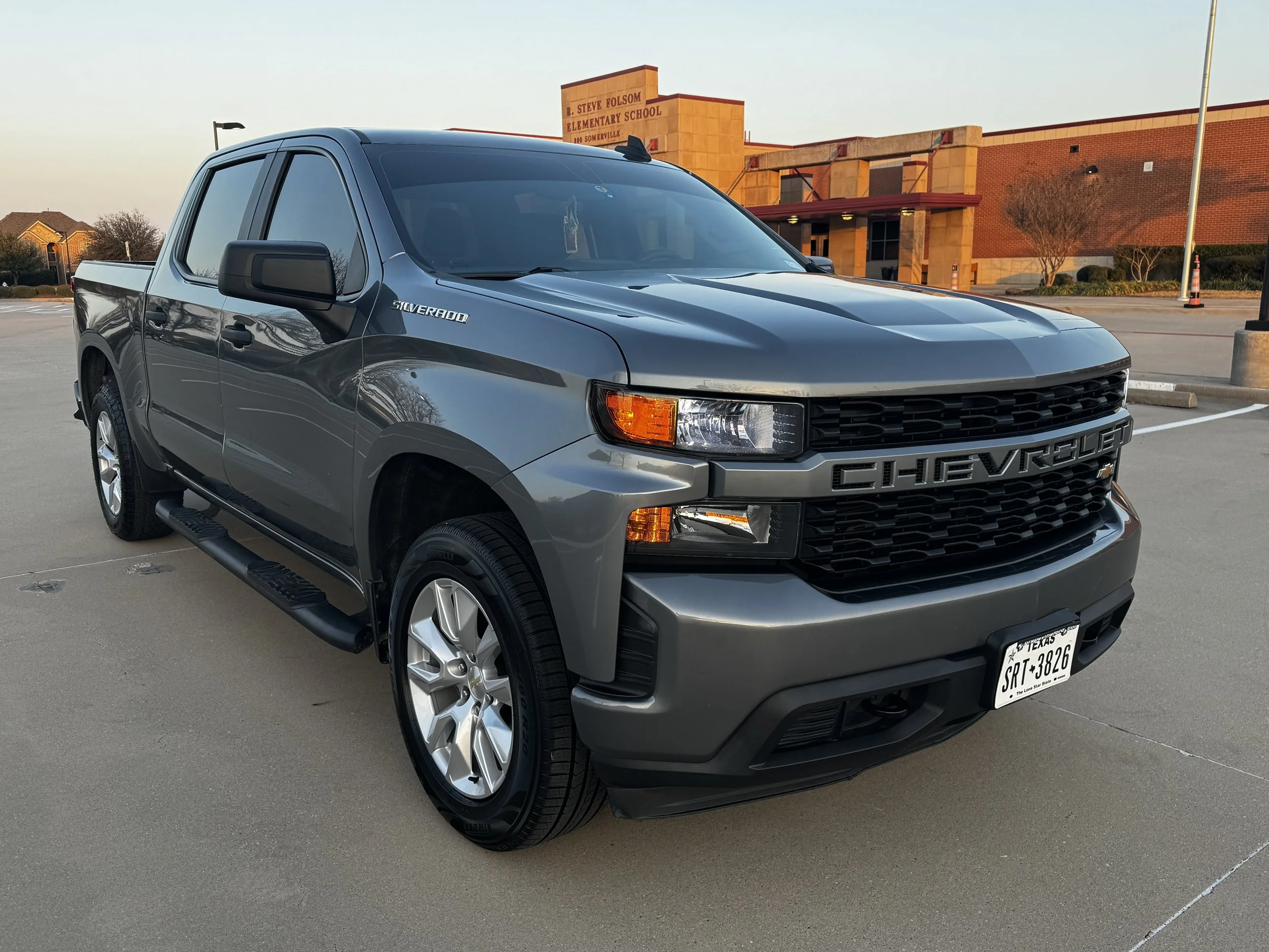 Gray Chevrolet Silverado pickup truck parked in a parking lot during sunset, with the elementary school building in the background.