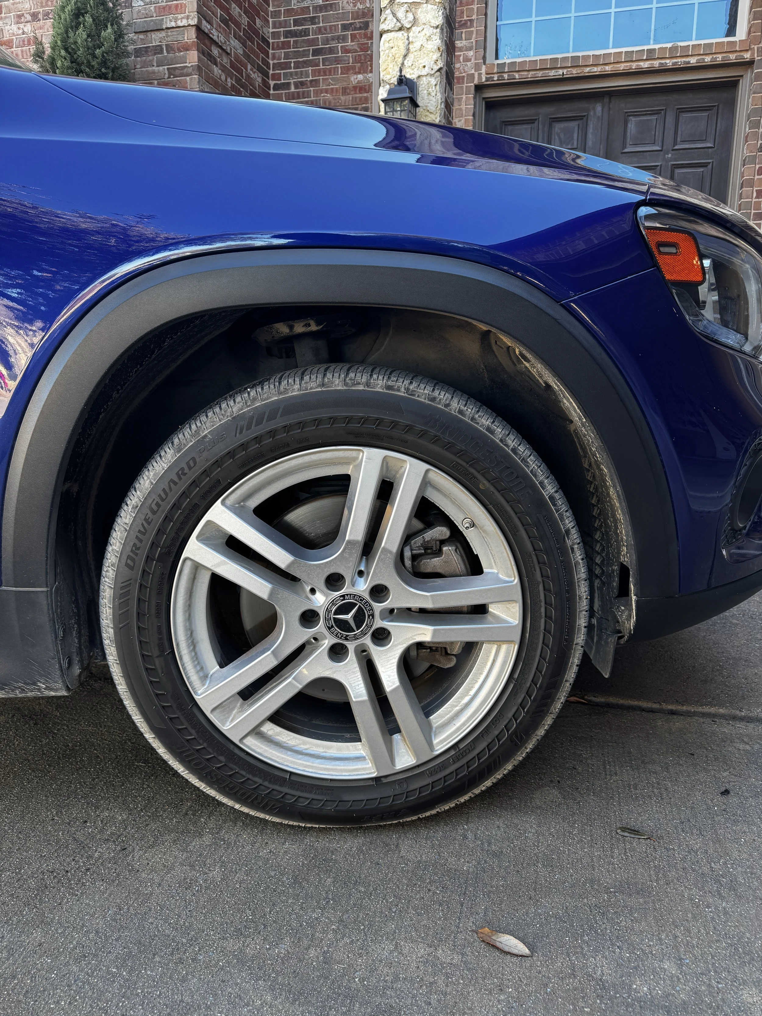 Close-up of a blue Mercedes-Benz vehicle's front wheel, tire, and fender, parked in front of a brick house.