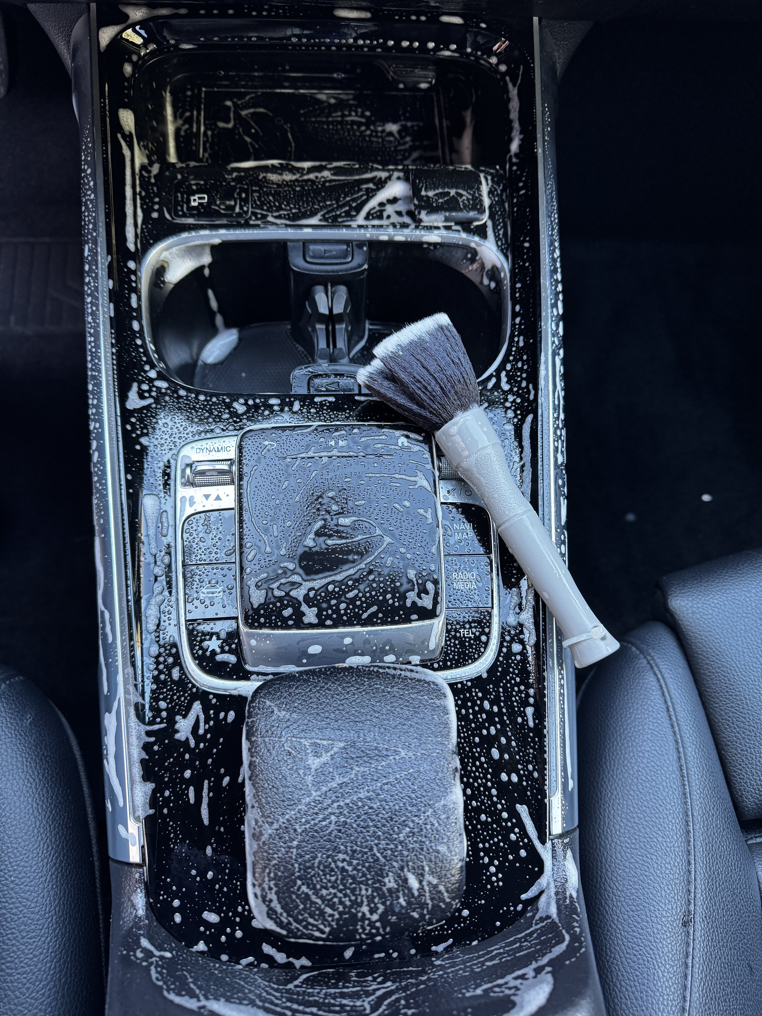 Car center console with spilled soap, a cleaning brush, and foam suds covering the surface and surrounding area.
