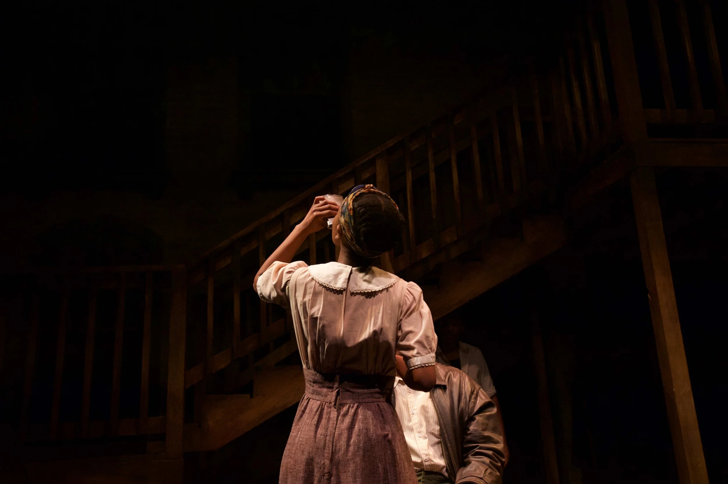 Theatre performance still of Ashli Rene Funches wearing a headscarf during a dramatic scene near a staircase on stage.
