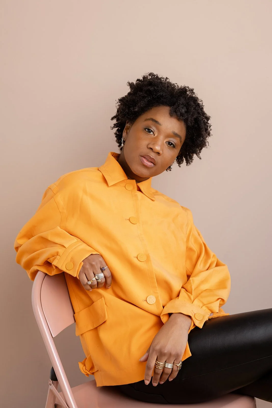 Studio portrait of Ashli Rene Funches wearing a yellow jacket and black pants, seated on a light pink chair against a beige background.