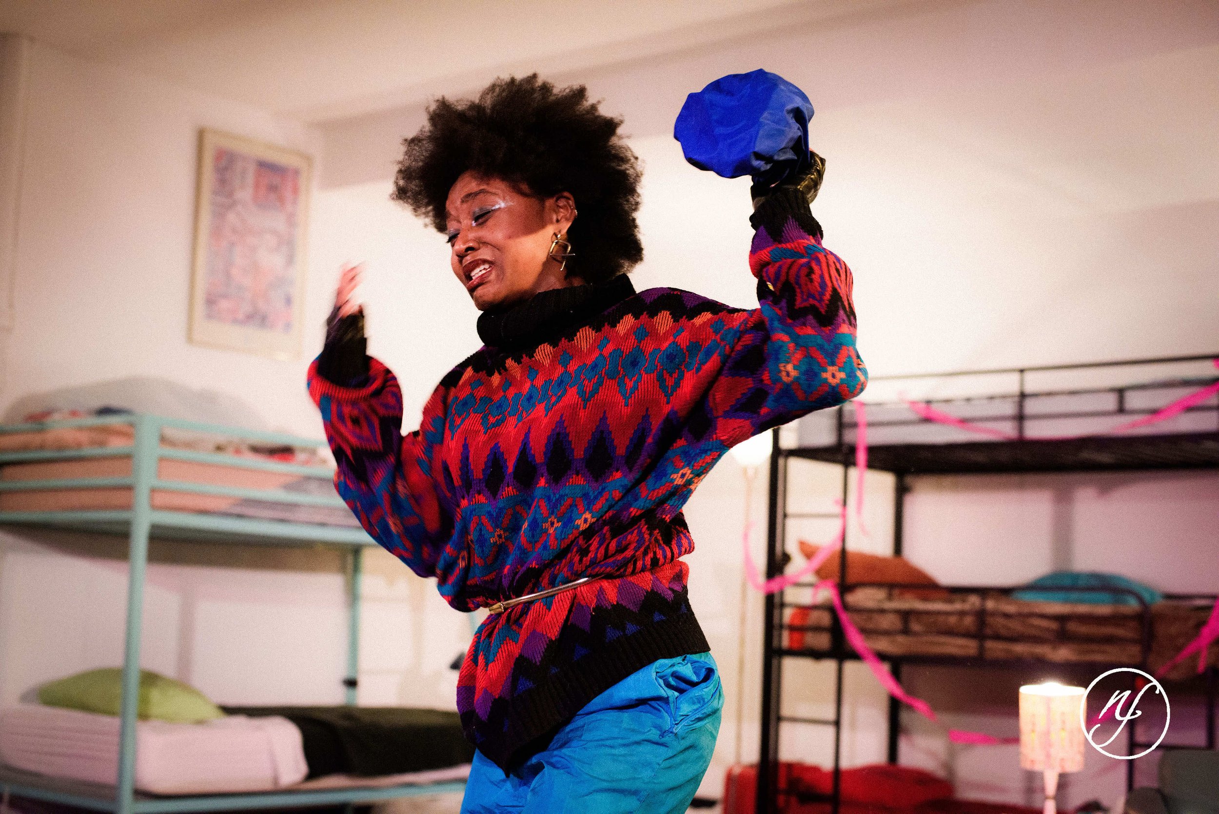 Theatre performance still of Ashli Rene Funches dancing in a bunk room set, wearing a patterned sweater and blue pants during a staged scene.