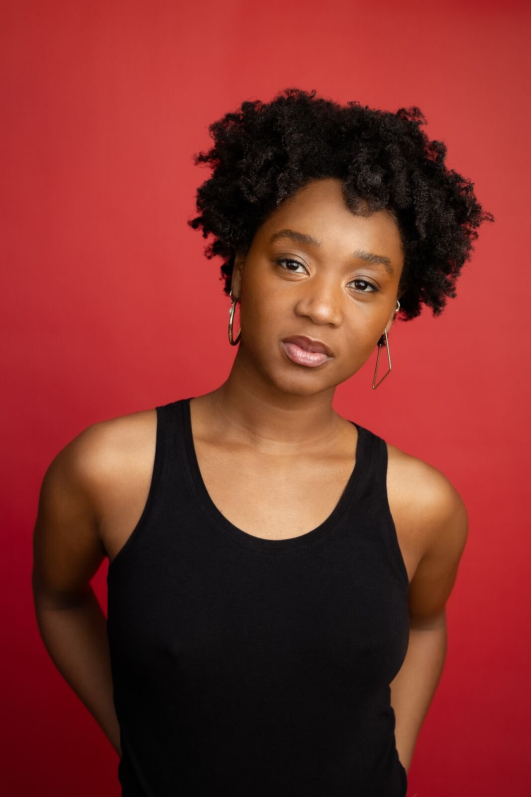 Studio portrait of Ashli Rene Funches with natural curly hair, wearing hoop earrings and a black sleeveless top against a red background.