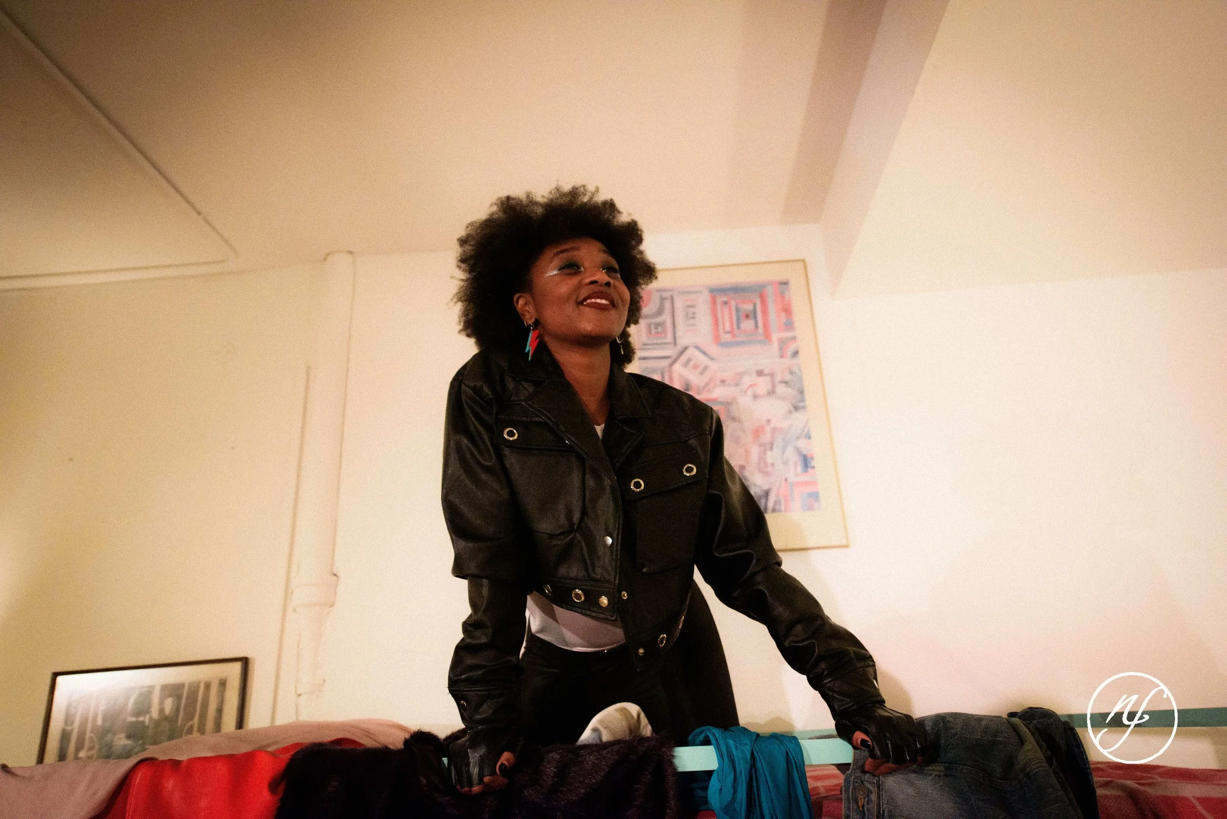 Theatre performance still of Ashli Rene Funches standing on a sofa and leaning toward a clothing rack during a staged scene.