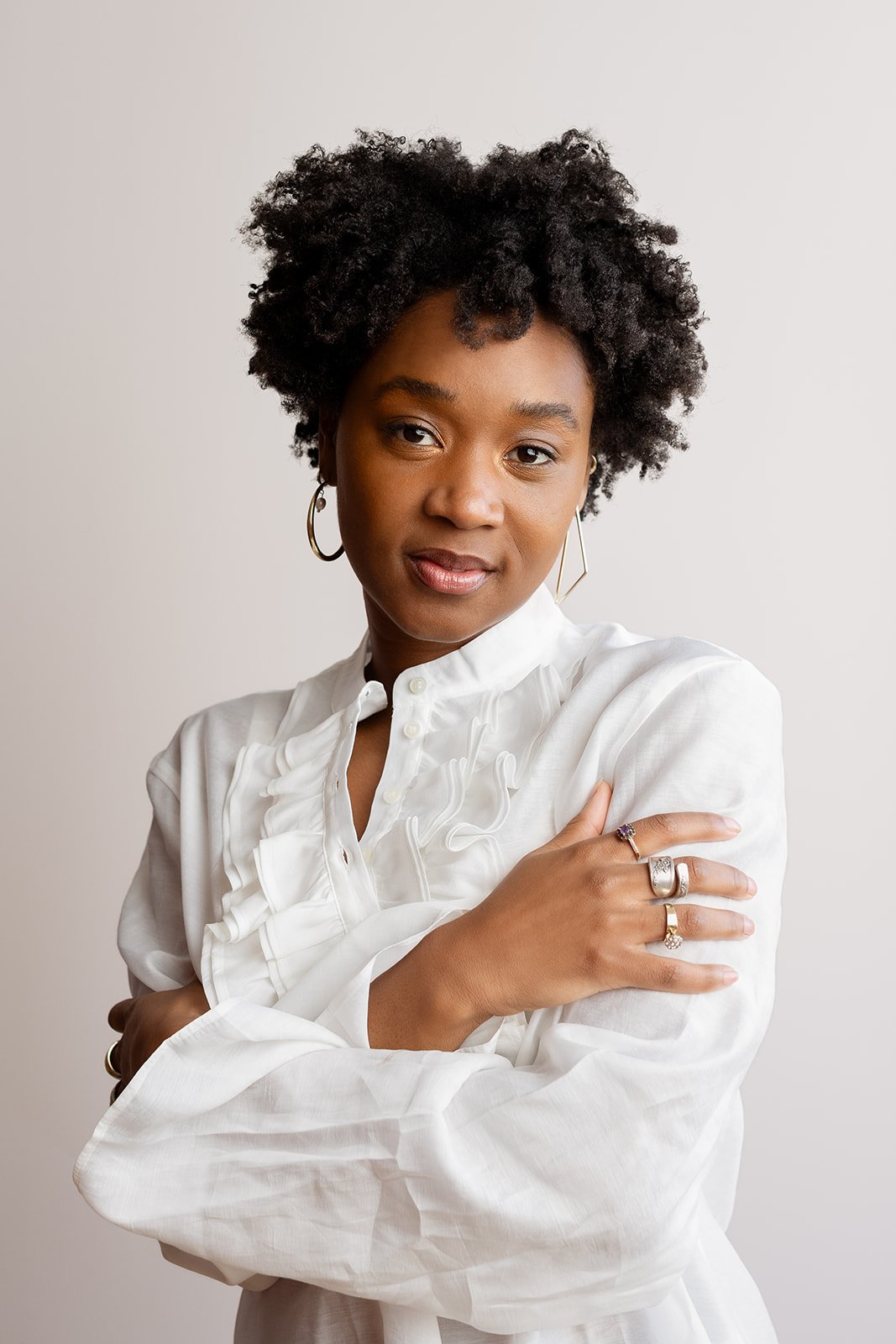 Professional headshot of Ashli Rene Funches with natural curly hair, wearing a white blouse and jewelry against a neutral studio background.