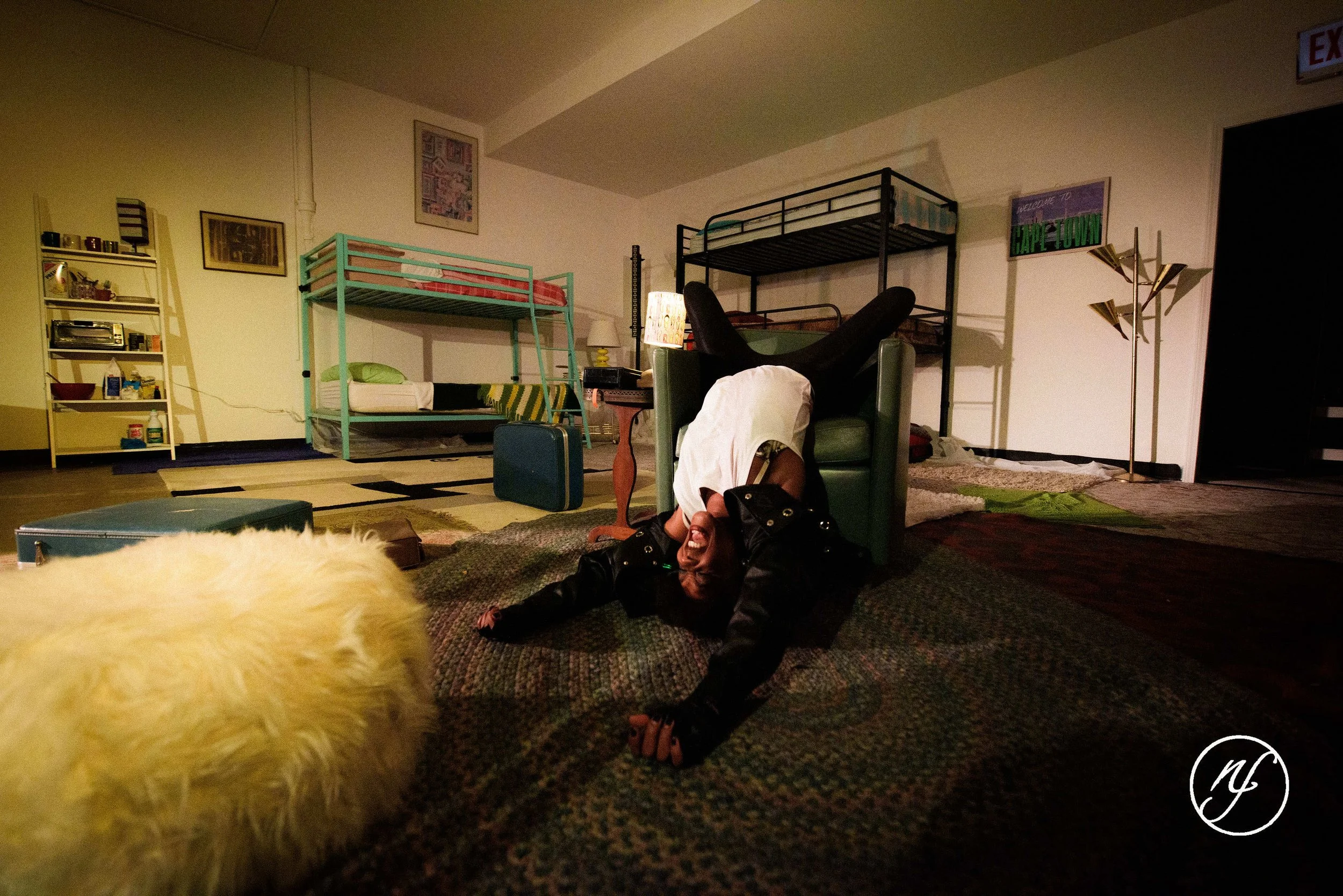Theatre performance still of Ashli Rene Funches laughing while leaning back in a green armchair within a cluttered stage set.