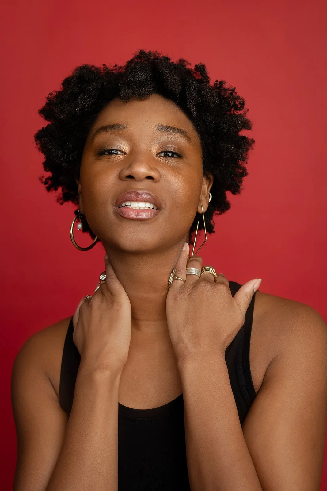 Studio headshot of Ashli Rene Funches with natural curly hair, wearing hoop earrings and a black sleeveless top against a red background.