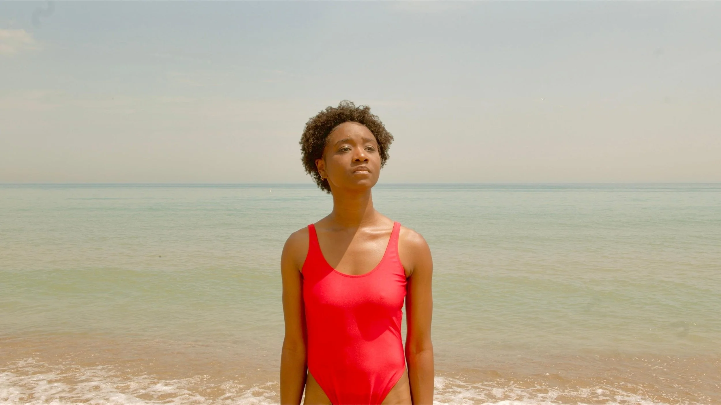 Performance still of Ashli Rene Funches in the film "A Yellow Circus", standing on a sandy beach in a red swimsuit with ocean water in the background.