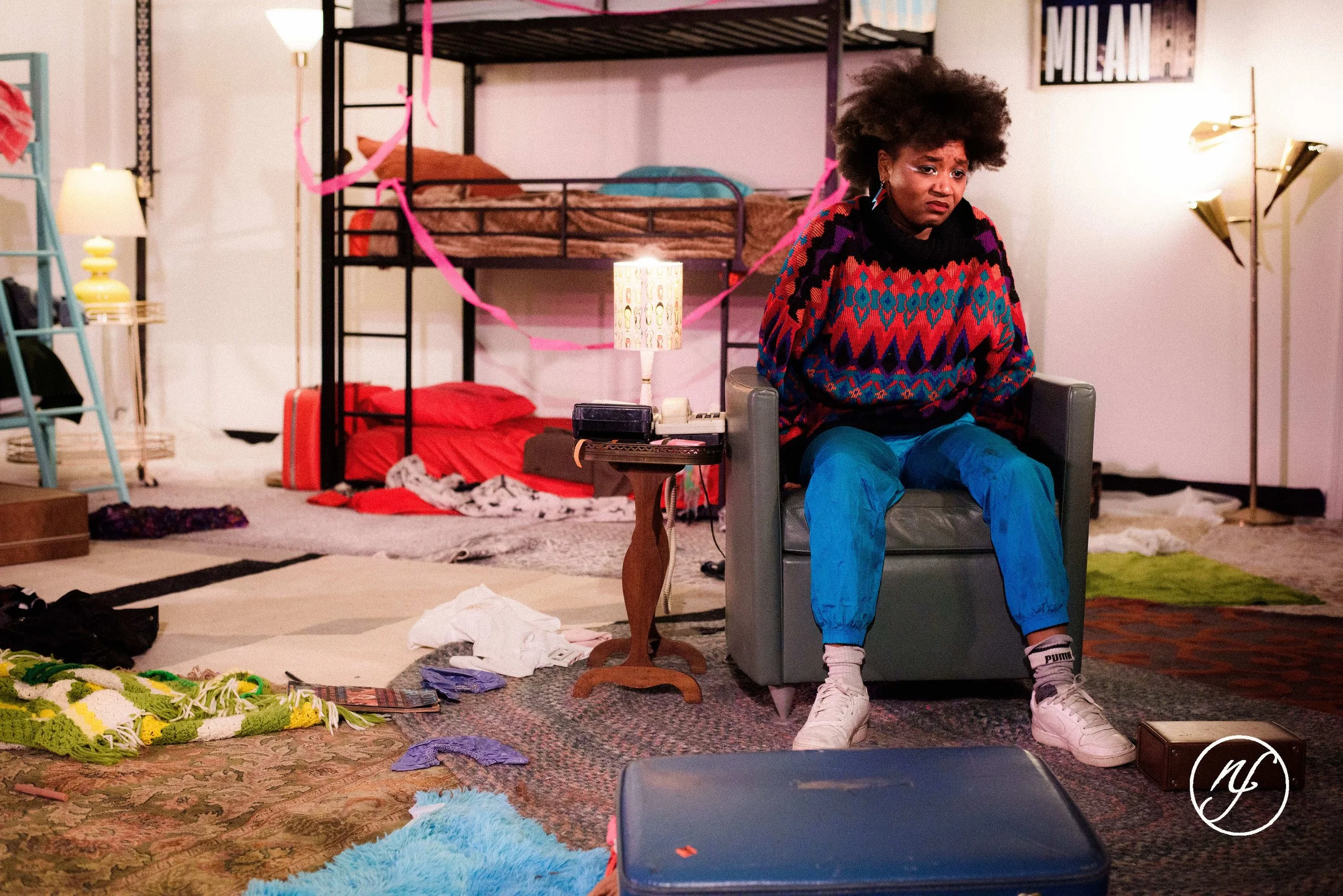 Theatre performance still of Ashli Rene Funches sitting in a gray armchair within a cluttered set during an emotional stage scene.