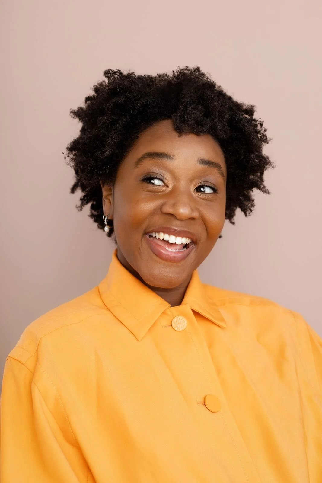 Professional headshot of Ashli Rene Funches with short natural curls, wearing a yellow button-up shirt against a light pink background.