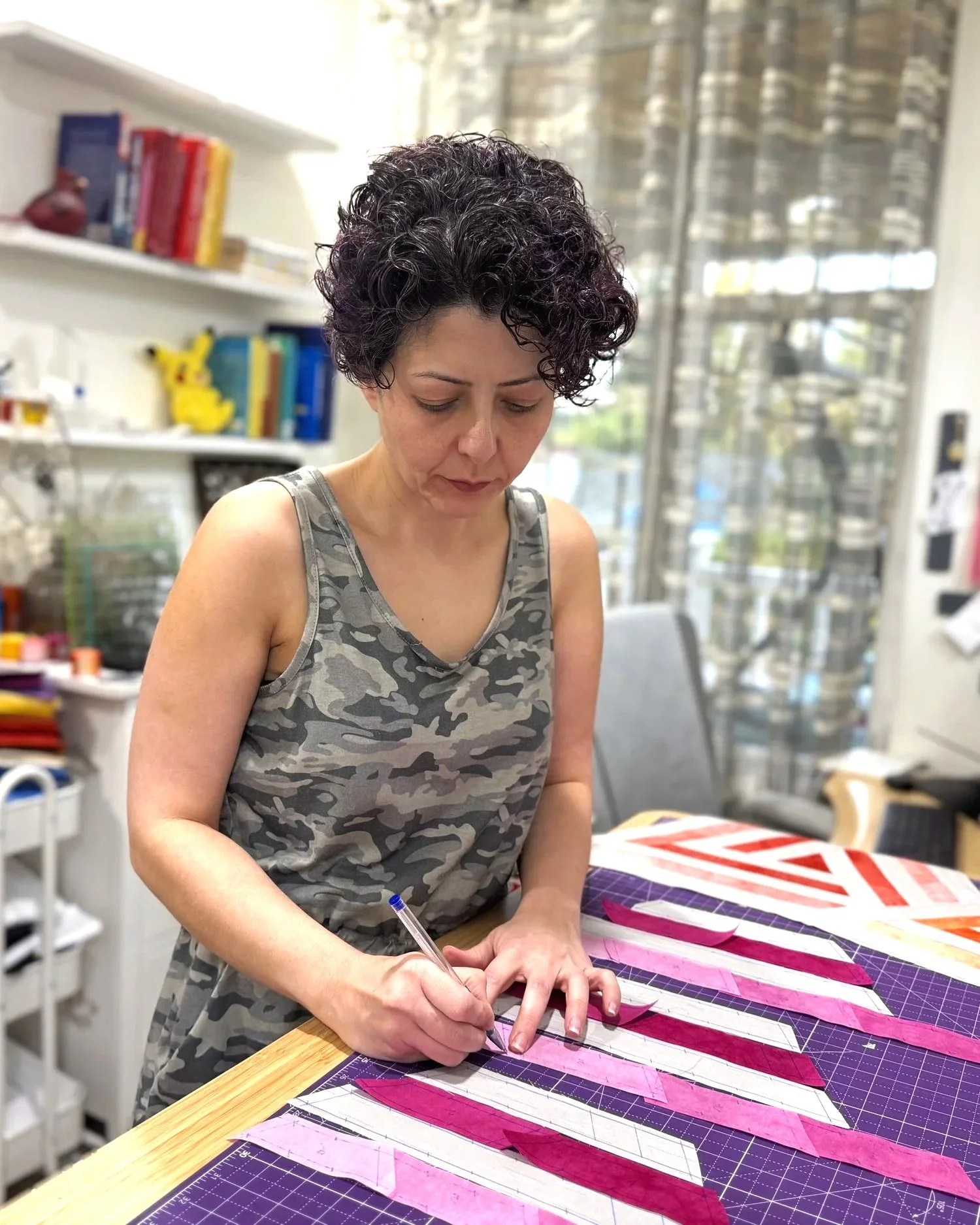A woman with short, curly dark hair wearing a gray camouflage tank top is working on a fabric project at a table, using a pen or marker to mark the fabric. The fabric pieces are arranged in a pattern with pink and white sections on a cutting mat. The background shows books and toys on shelves, a window with blinds, and a cozy workshop setting.