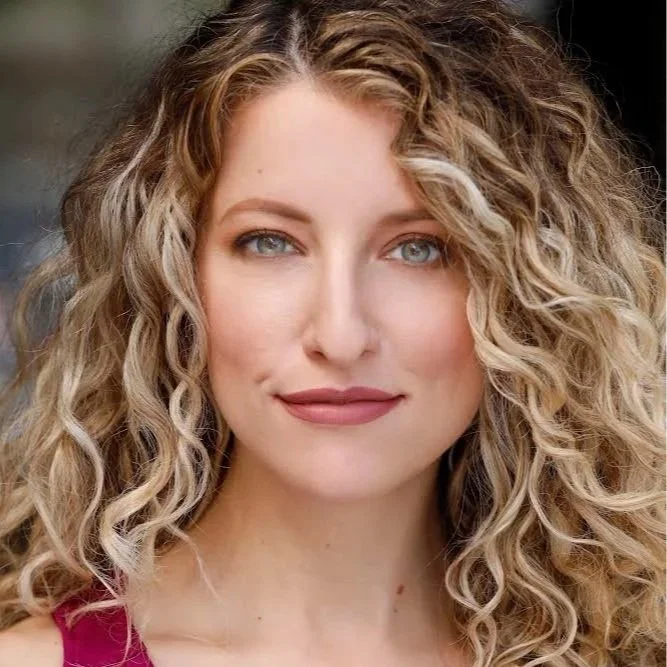 Portrait of a young woman with long, dark brown hair, fair skin, and makeup, smiling softly, against a plain gray background.