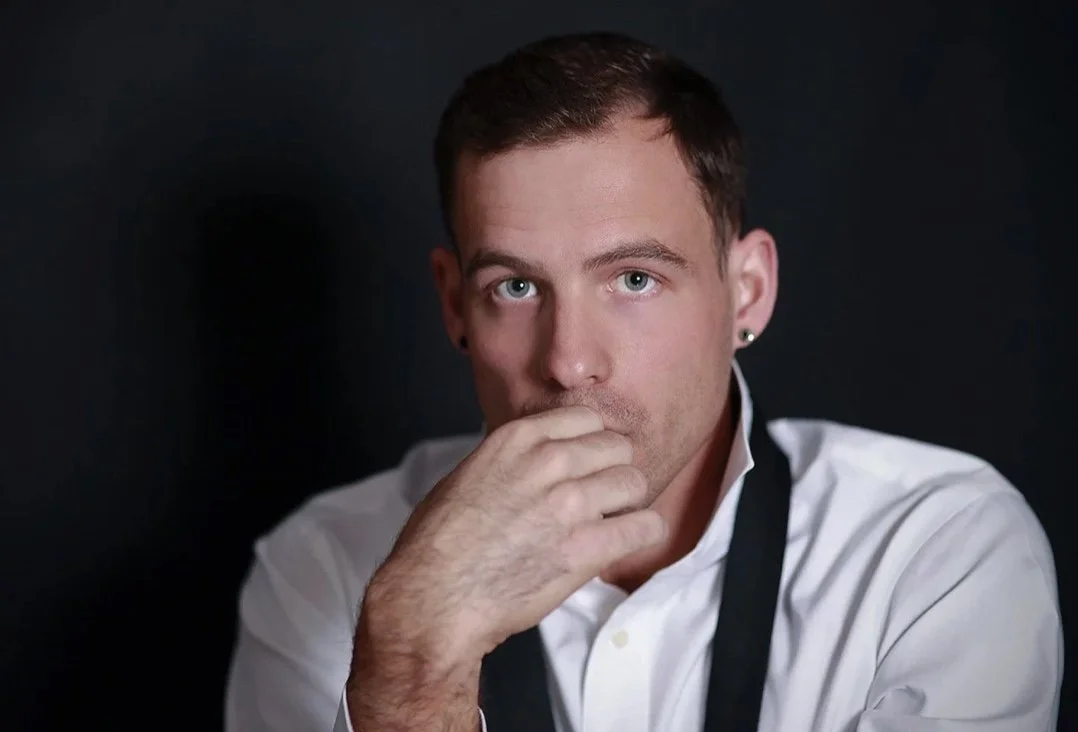A man with short brown hair and earrings, wearing a white shirt and black apron, looks directly at the camera with his hand partially covering his mouth against a black background.